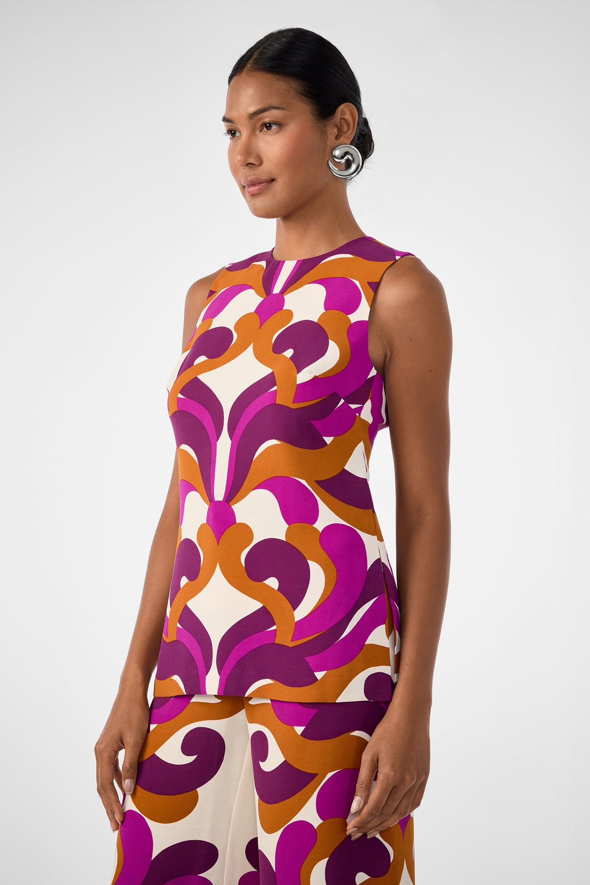 A woman with dark hair pulled back wears the Haruto Top, a sleeveless piece with a bold purple, orange, and cream pattern. She stands against a plain light background, looking slightly to the side.