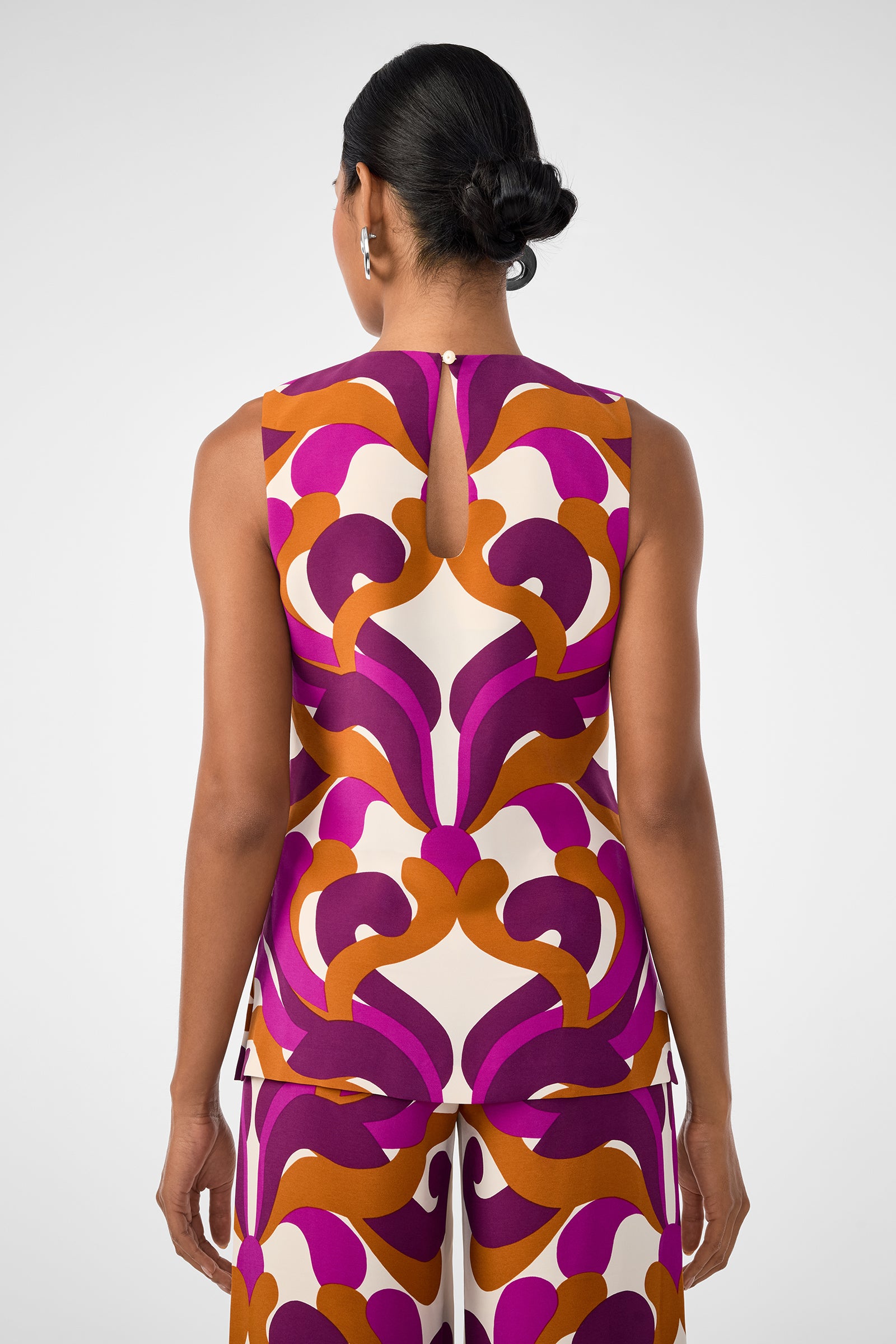 A woman with dark hair in a low bun is seen from behind, wearing the sleeveless Haruto Top and matching pants featuring a bold geometric pattern in purple, orange, and cream.