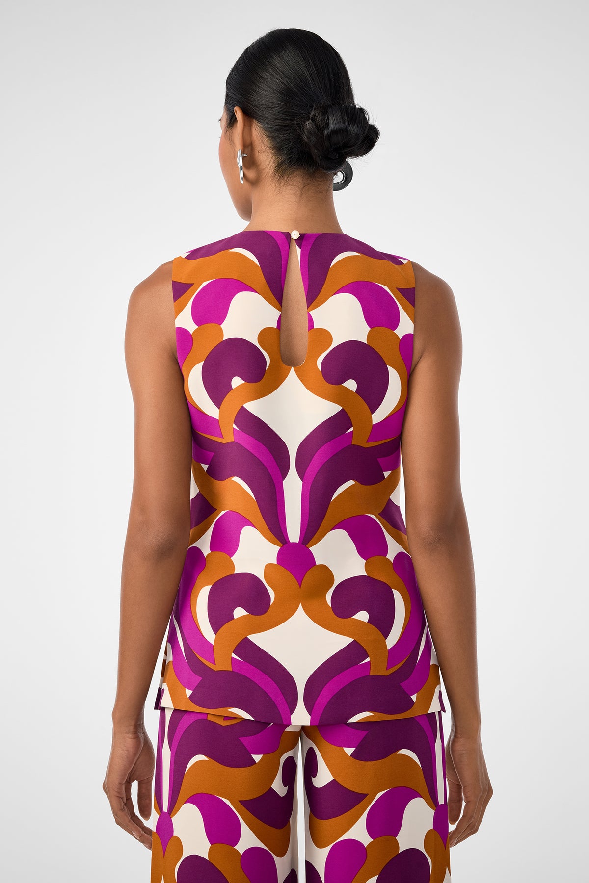 A woman with dark hair in a low bun is seen from behind, wearing the sleeveless Haruto Top and matching pants featuring a bold geometric pattern in purple, orange, and cream.