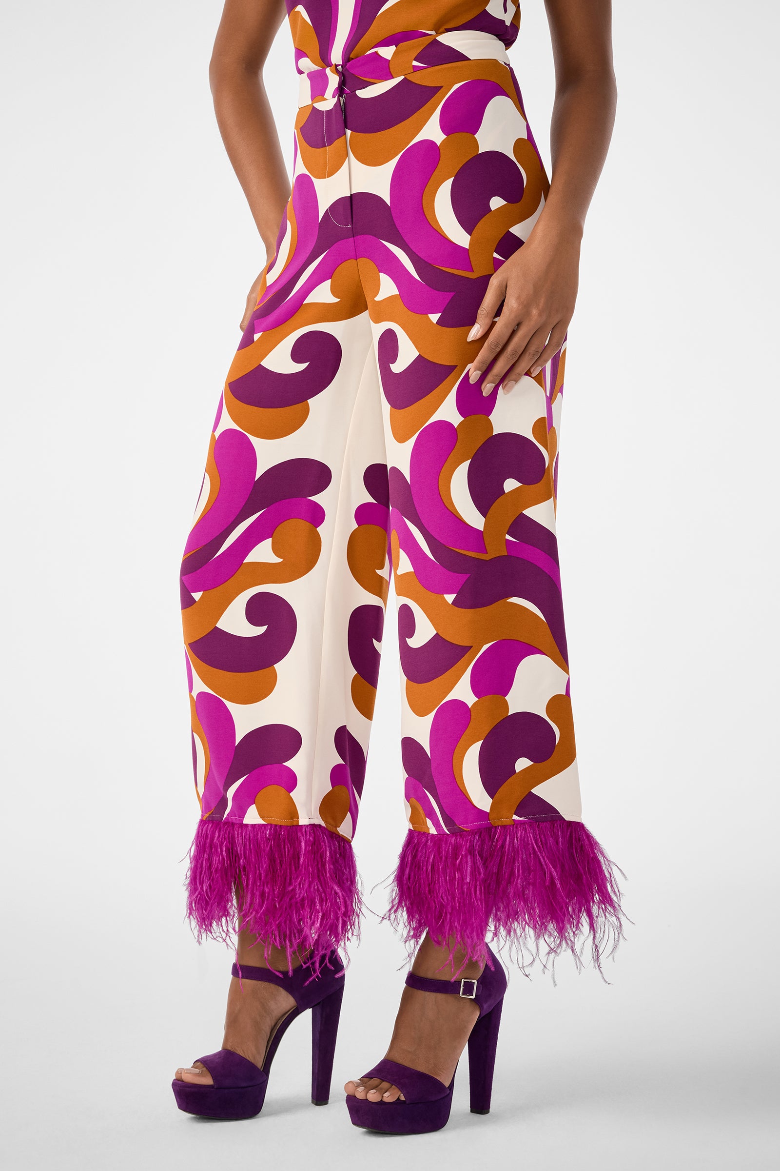 Wearing the Short Weekend Pant, featuring high-waisted, wide-leg style with a vibrant orange, purple, and pink swirl pattern and purple feather-trimmed hems, paired with purple platform heels.