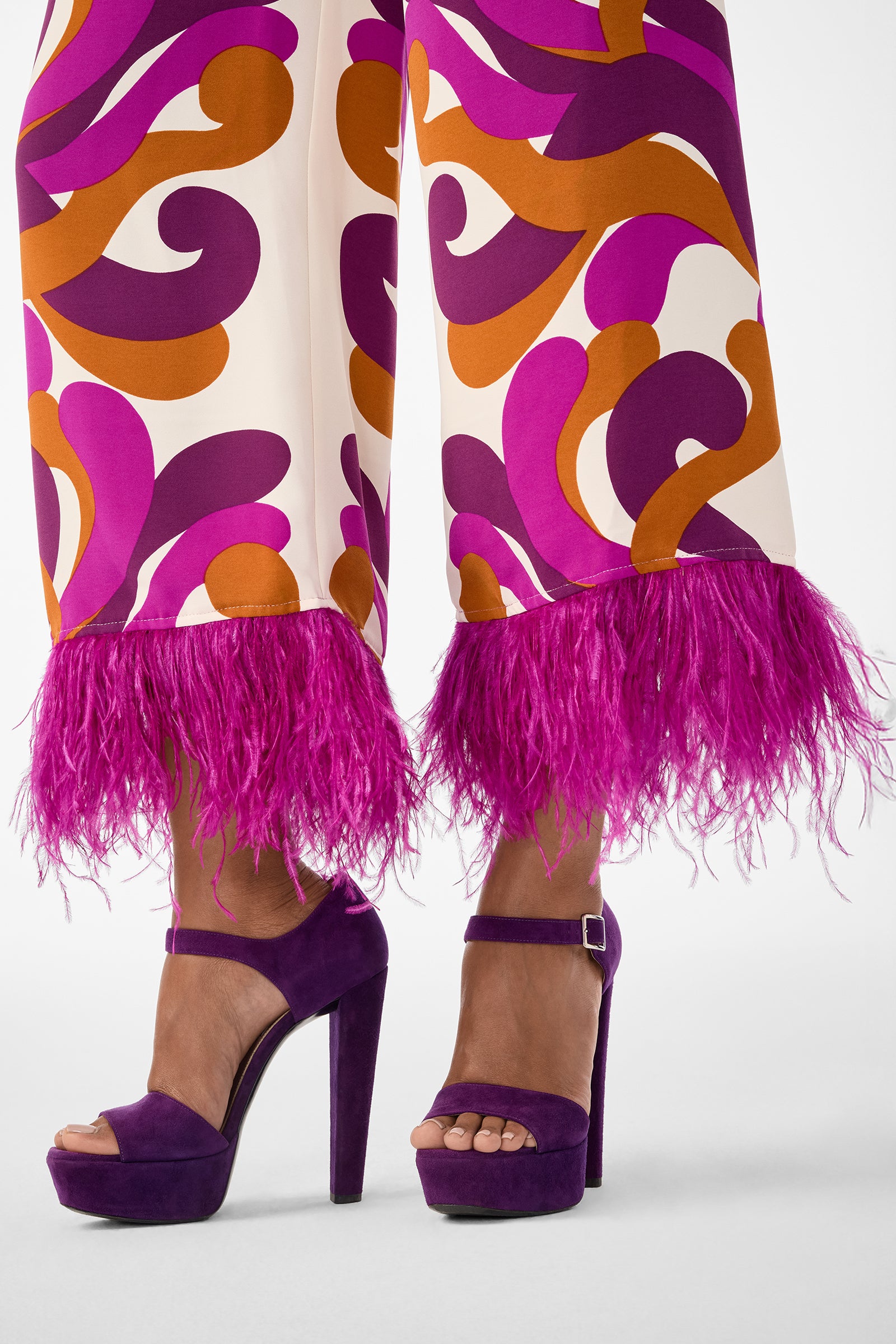 Close-up of a person wearing the Short Weekend Pant, featuring orange and purple abstract patterns with bright pink feathered cuffs, paired with high-heeled purple suede platform sandals.
