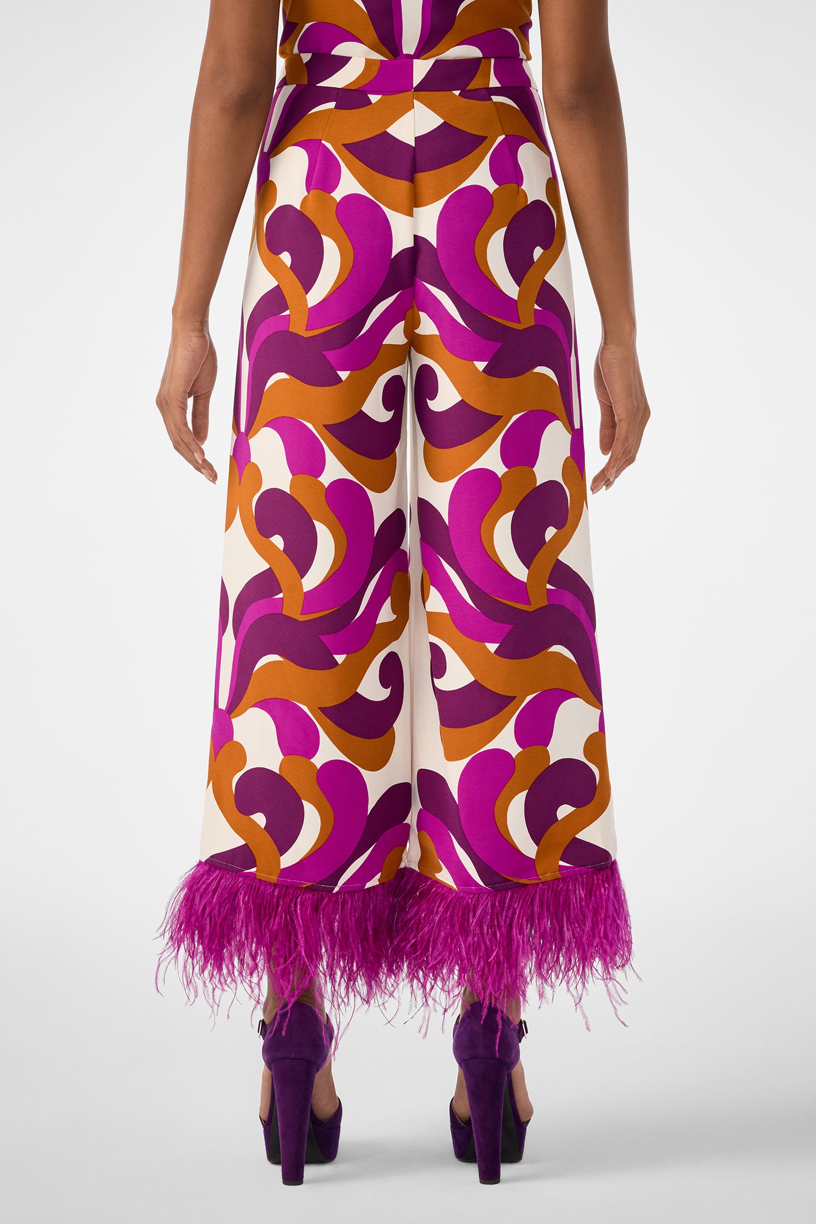 A person faces away, wearing the Short Weekend Pant—high-waisted, wide-leg with bold purple, orange, and cream swirls and purple feather hem—paired with purple high heels.