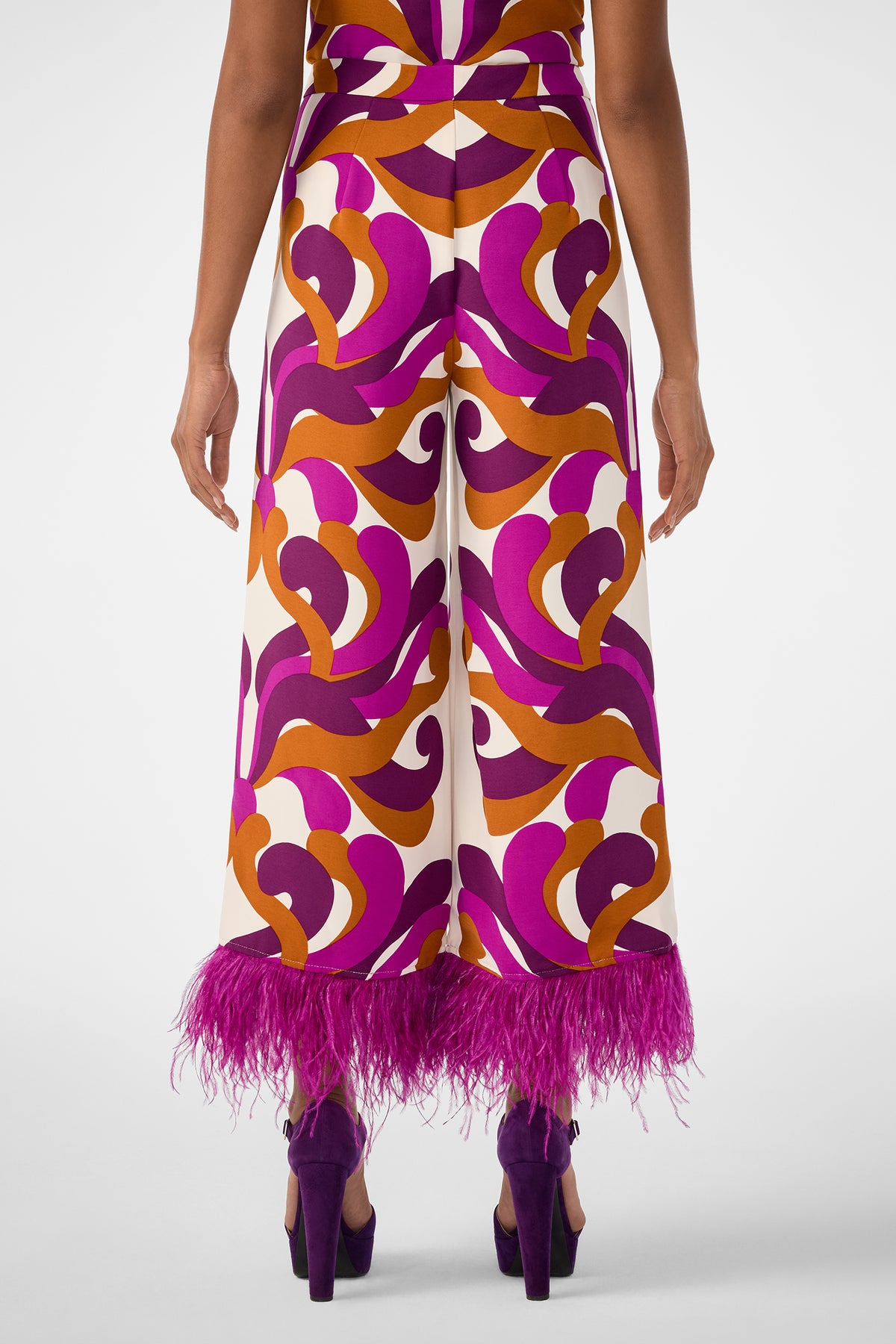 A person faces away, wearing the Short Weekend Pant—high-waisted, wide-leg with bold purple, orange, and cream swirls and purple feather hem—paired with purple high heels.