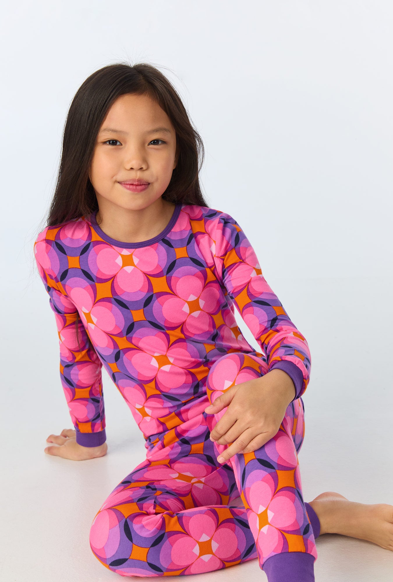 A young girl with long dark hair sits on the floor, smiling slightly. She wears the Space Cat Kids Long-Sleeve Long Pant Jersey PJ Set in multi-colored hues. The background is plain white.