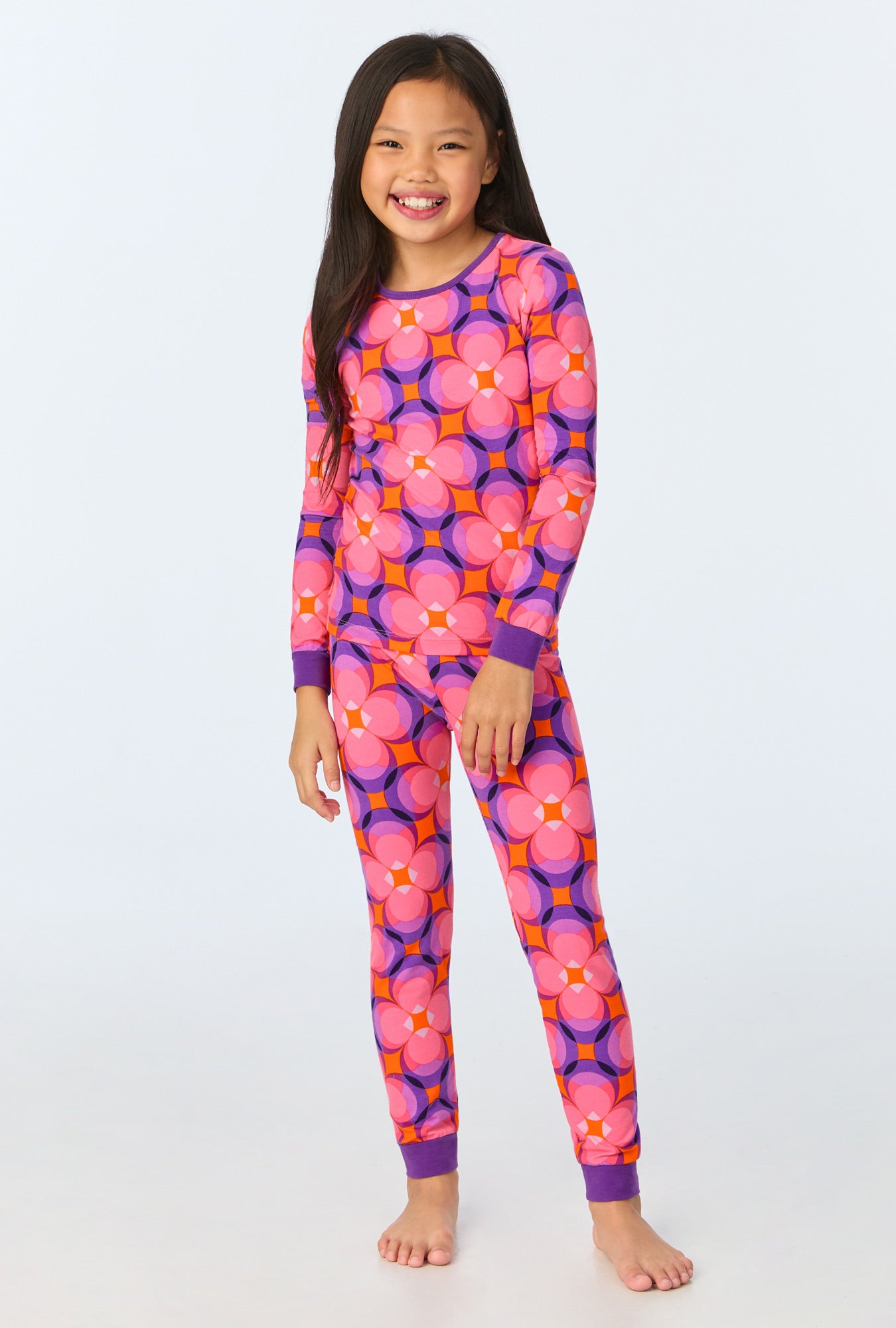 A smiling young girl with long dark hair wears the Space Cat Kids Long-Sleeve Long Pant Jersey PJ Set, standing barefoot against a plain light background.