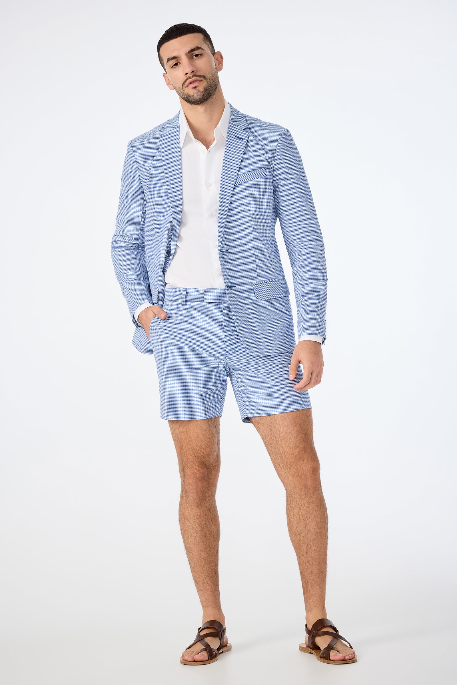 A man poses confidently in the Roger Short - Ocean Gingham set: a light blue seersucker suit with matching shorts, paired with a white dress shirt and brown sandals, against a plain white background.