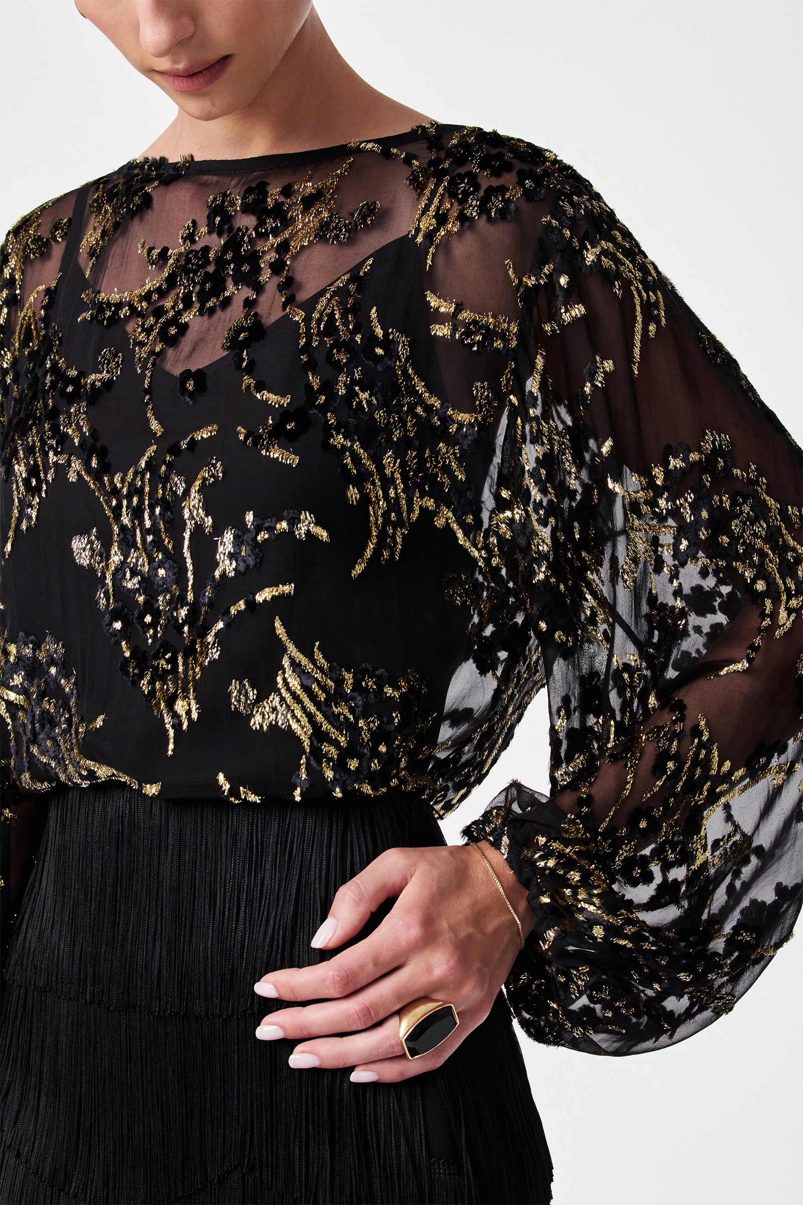 A woman wears the Ursa Metallic Burnout Balloon Sleeve Top with sheer black fabric, gold and black sequin embroidery, voluminous sleeves, and a large statement ring. Her manicured hand rests on her hip.