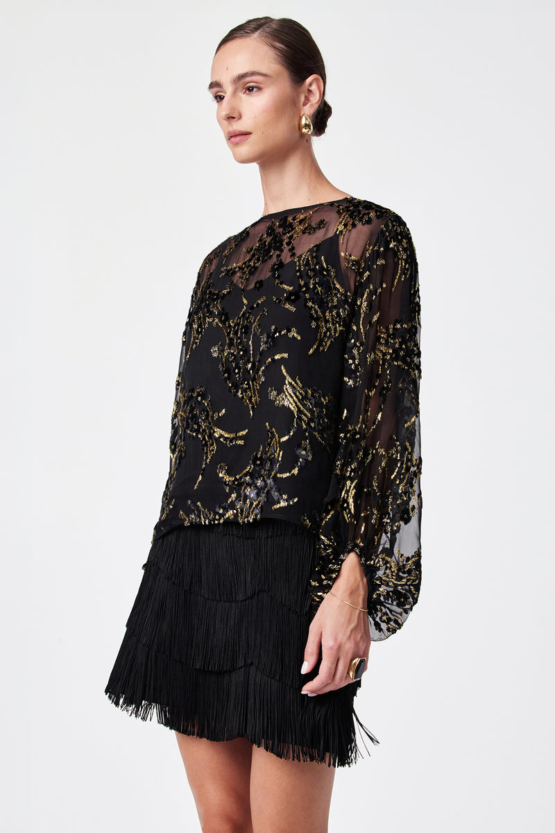 Wearing the Ursa Metallic Burnout Balloon Sleeve Top—a sheer black top with gold sequin patterns—paired with a black fringed mini skirt, gold hoop earrings, and a gold ring, against a plain white background.