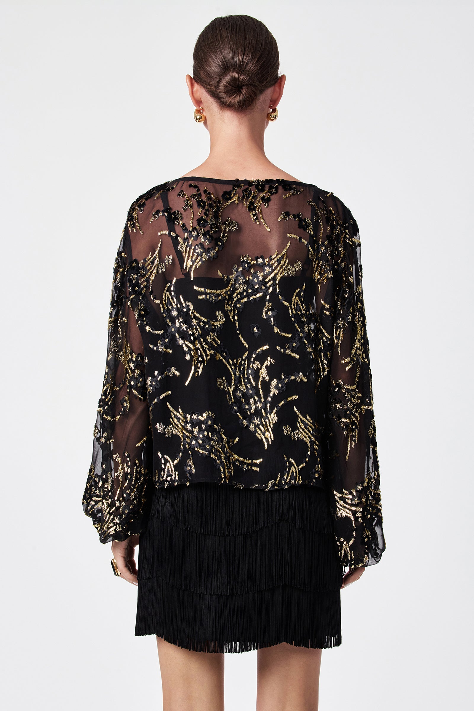 A woman with her hair in a bun is shown from the back, wearing the Ursa Metallic Burnout Balloon Sleeve Top and a short black fringed skirt, accessorized with gold hoop earrings.