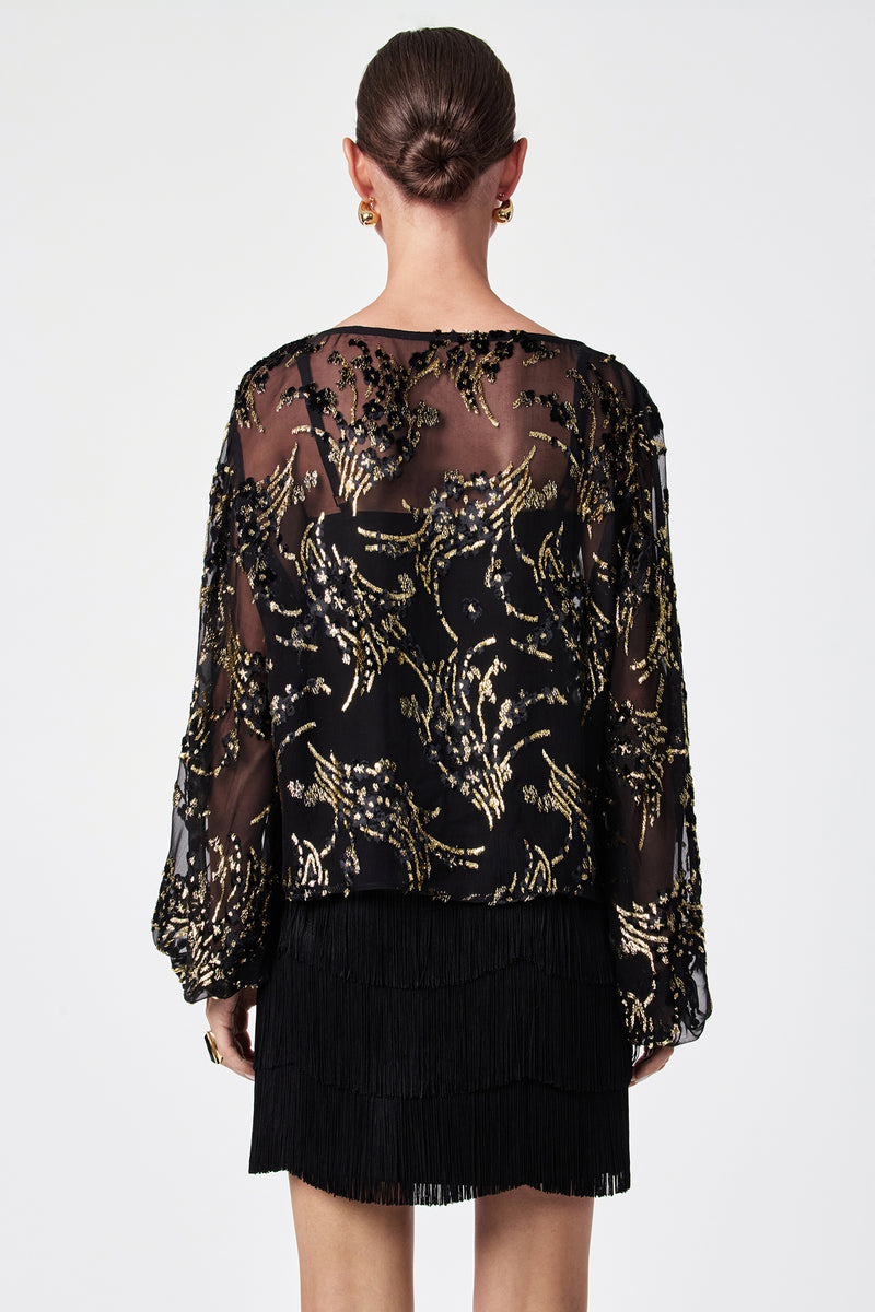 A woman with her hair in a bun is shown from the back, wearing the Ursa Metallic Burnout Balloon Sleeve Top and a short black fringed skirt, accessorized with gold hoop earrings.