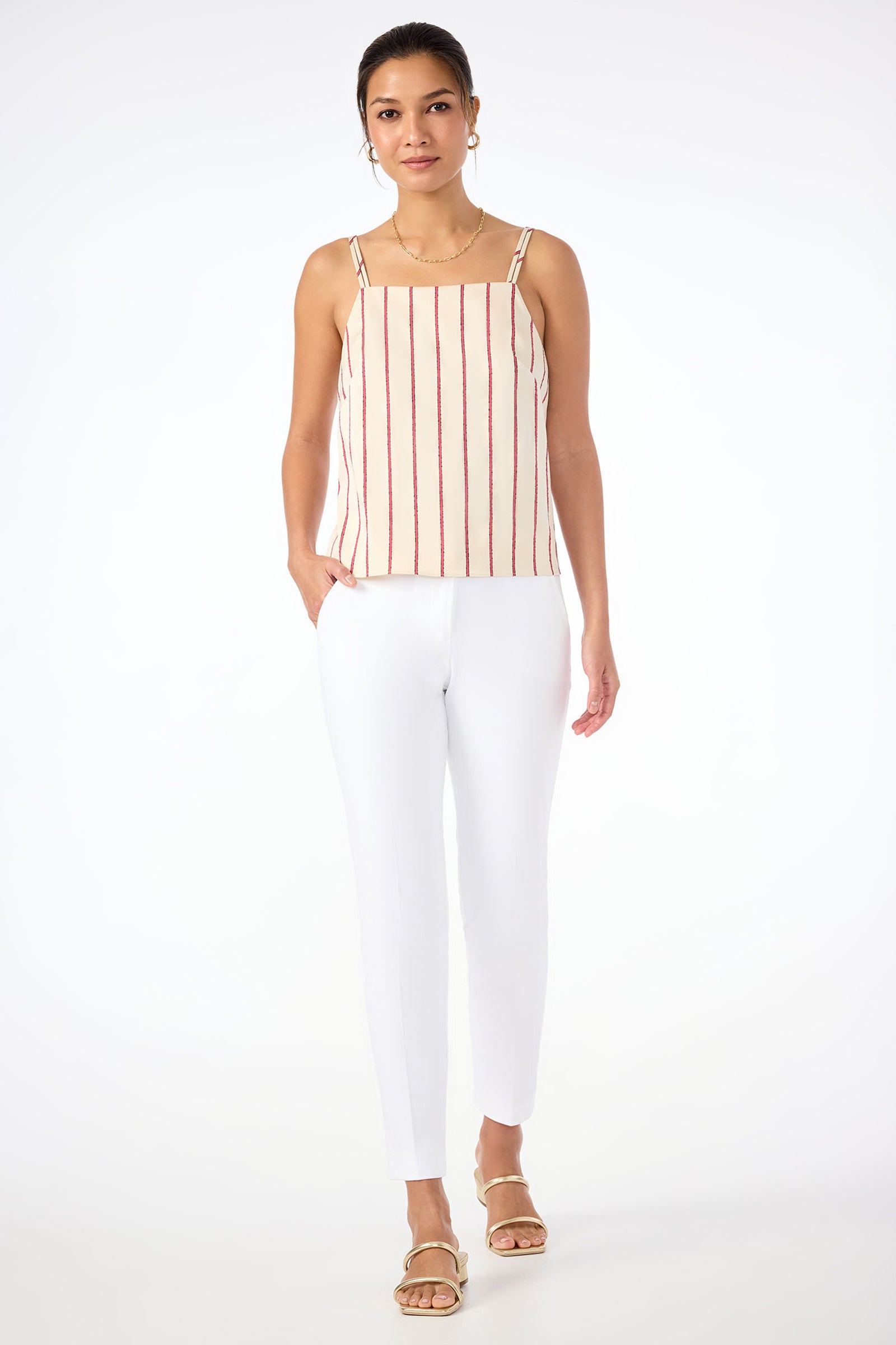 A woman stands against a white background wearing the Trouve Sleeveless A-Line Top in Solaire Stripe with white slim-fit pants, gold hoop earrings, and gold sandals. One hand is in her pocket as she looks forward.