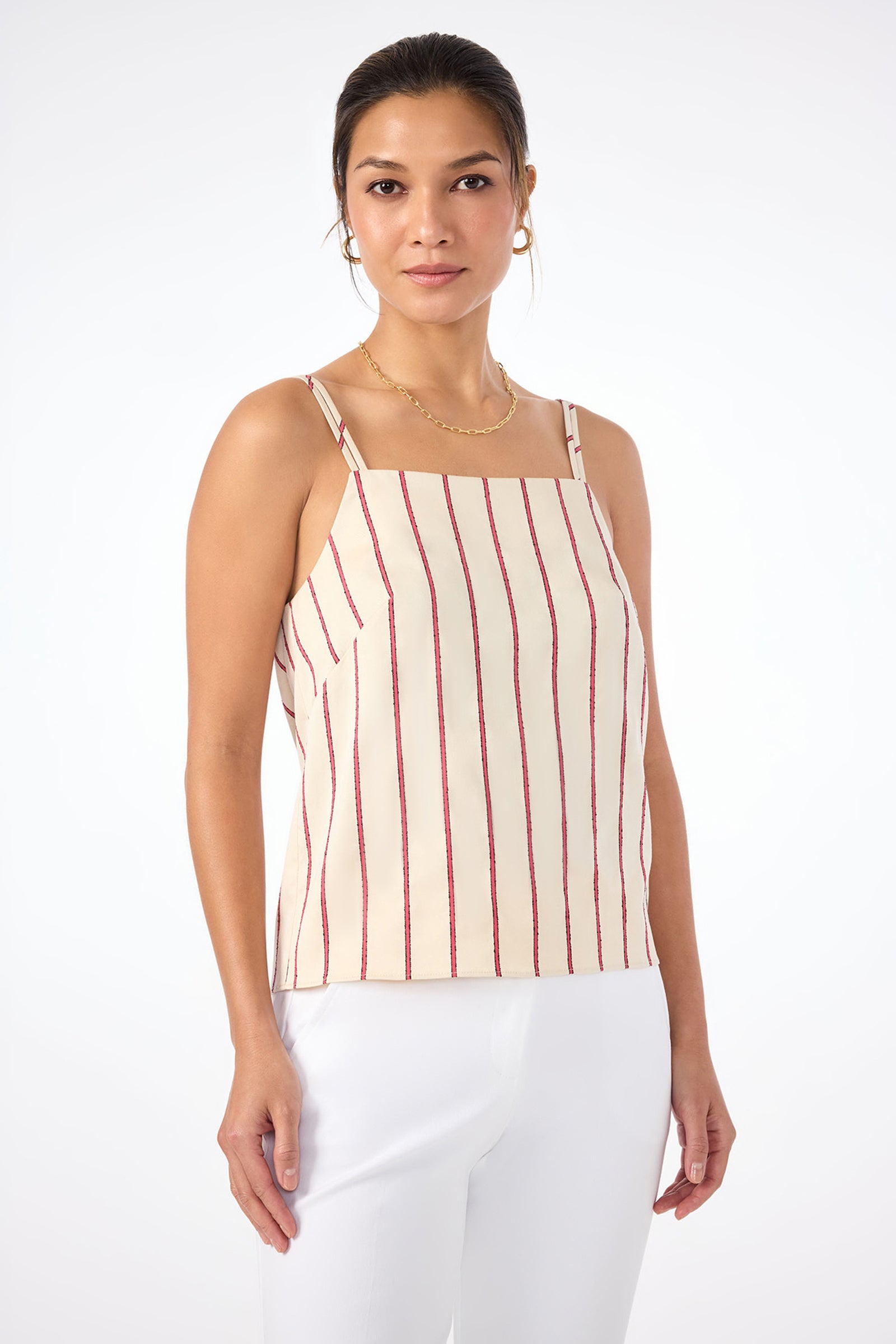 A woman stands against a white background wearing the Trouve Sleeveless A-Line Top in Solaire Stripe with white pants, her hair tied back, accessorized with gold hoop earrings and a necklace.