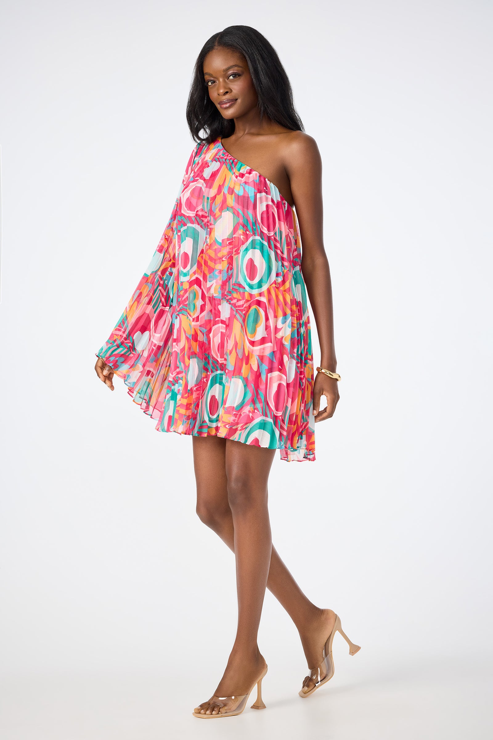 A woman models the Narbonne One-Shoulder Mini Dress - Dramont, featuring a vibrant geometric print and flowy mini silhouette. She pairs it with clear high heels and stands against a plain white background.