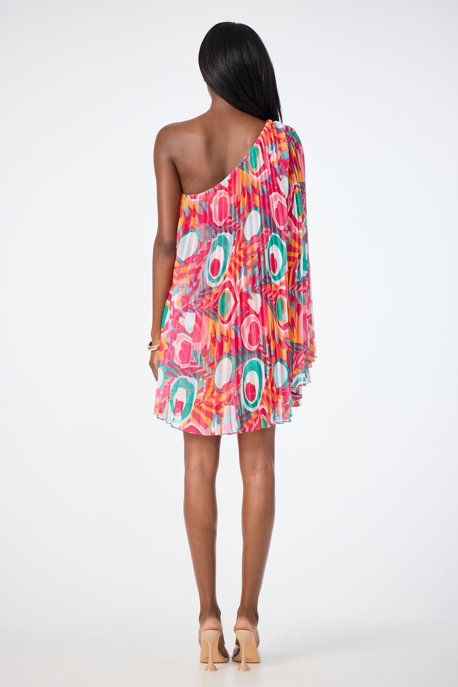 A woman with long dark hair and beige heels faces away, wearing the Narbonne One-Shoulder Mini Dress - Dramont, a colorful, above-the-knee dress featuring a bold geometric pattern in red, pink, green, and white against a plain white background.