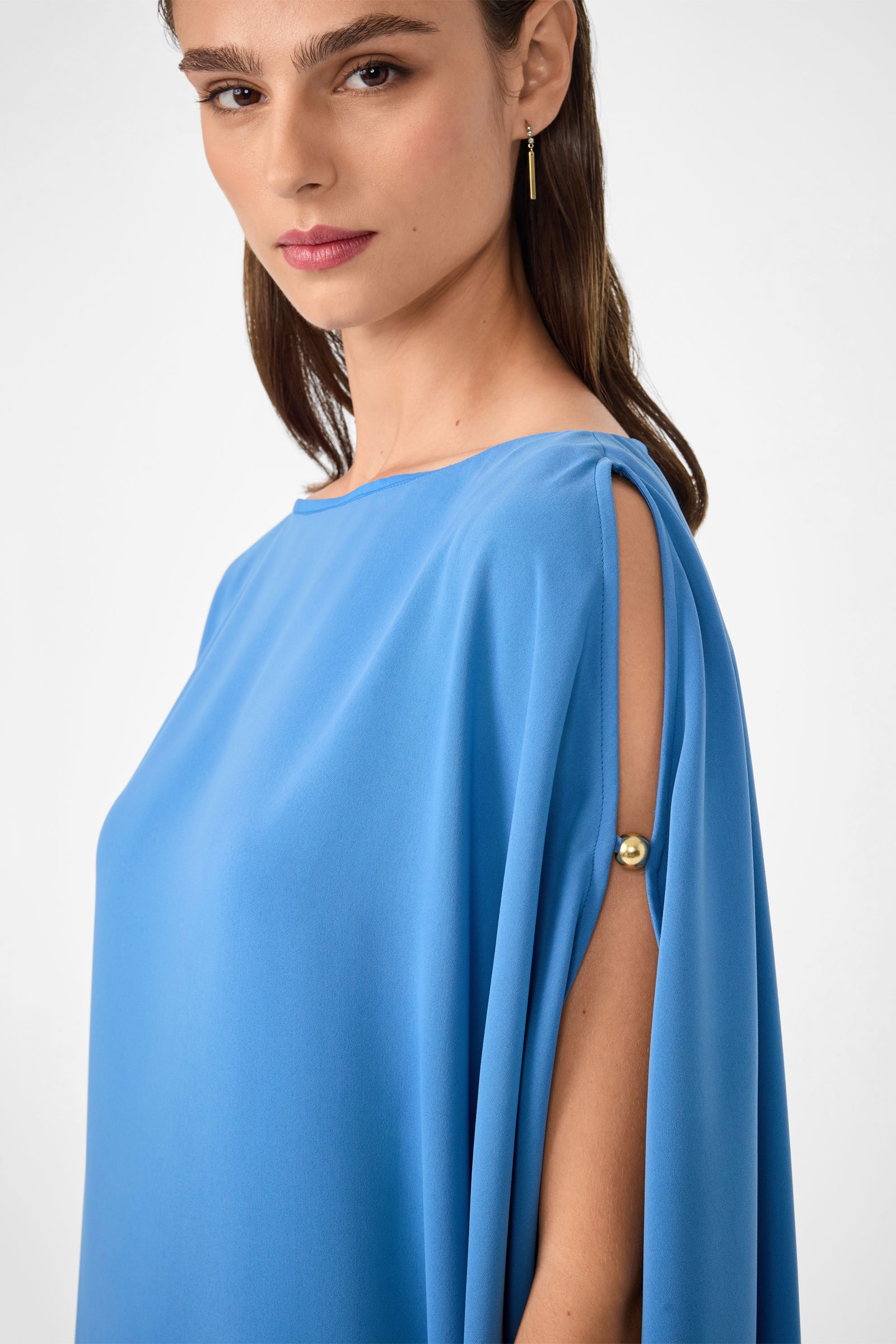 A woman stands against a plain white background wearing the Razu Short Split Sleeve Mini Dress, a loose-fitting, light blue dress with open slit sleeves fastened by gold buttons.