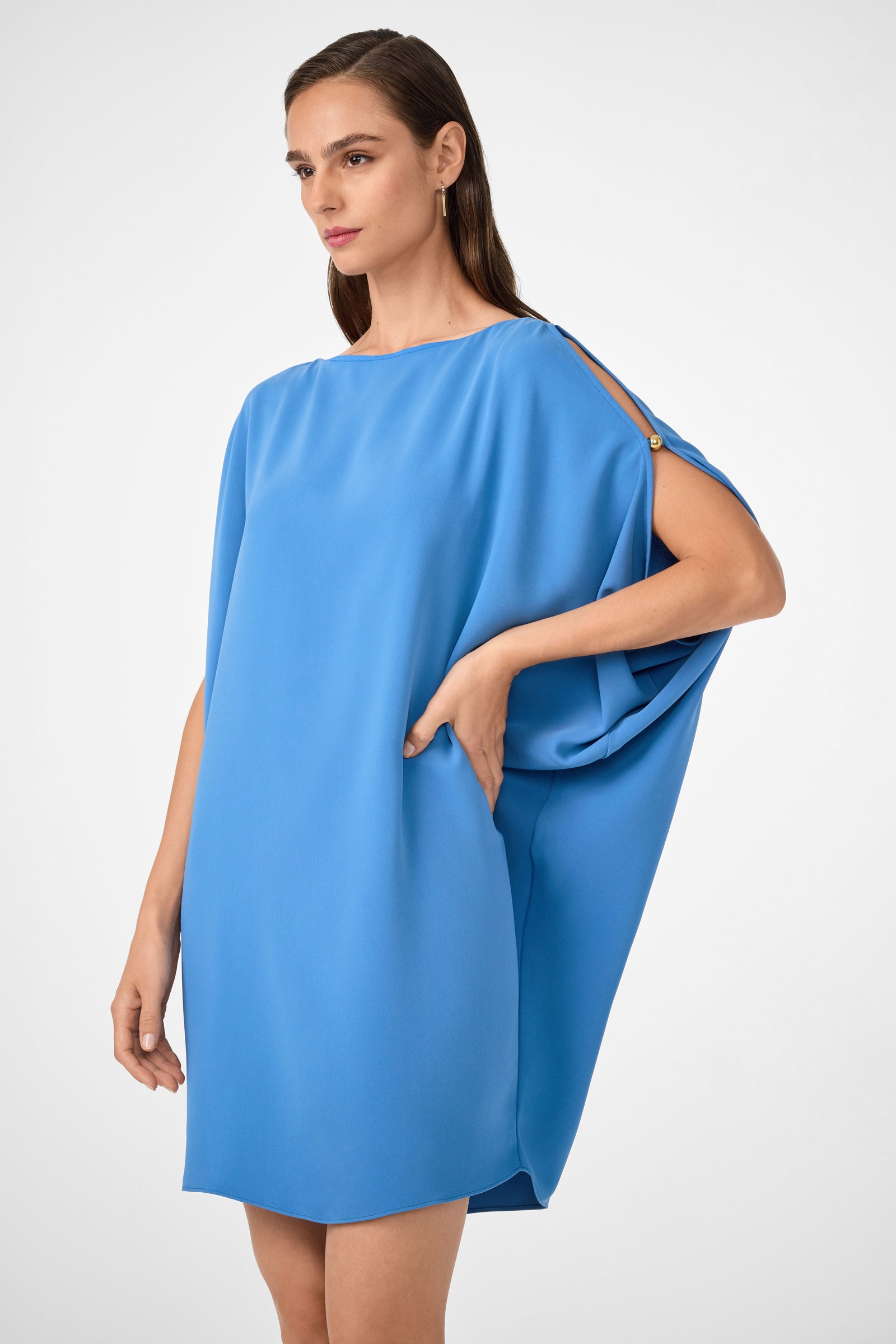 A woman poses with one hand on her hip against a plain light background, wearing the Razu Short Split Sleeve Mini Dress—loose-fitting, bright blue, featuring a draped one-shoulder sleeve and gold button detail.