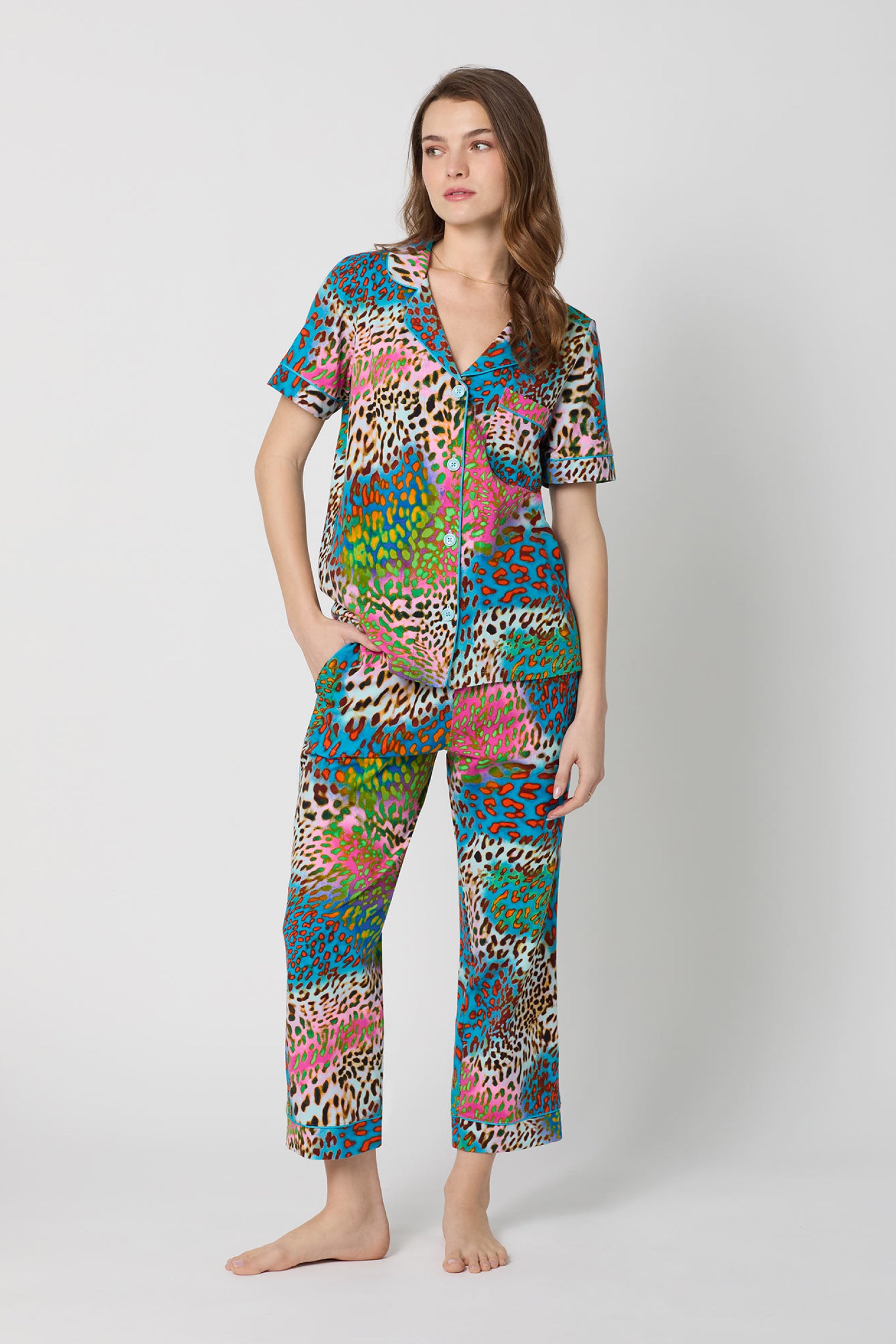 A woman with long brown hair stands barefoot against a plain background, wearing the Crop Classic PJ Set - Spray Leopard: a colorful, animal-print matching set featuring a short-sleeve button-up top and pants. She looks relaxed.