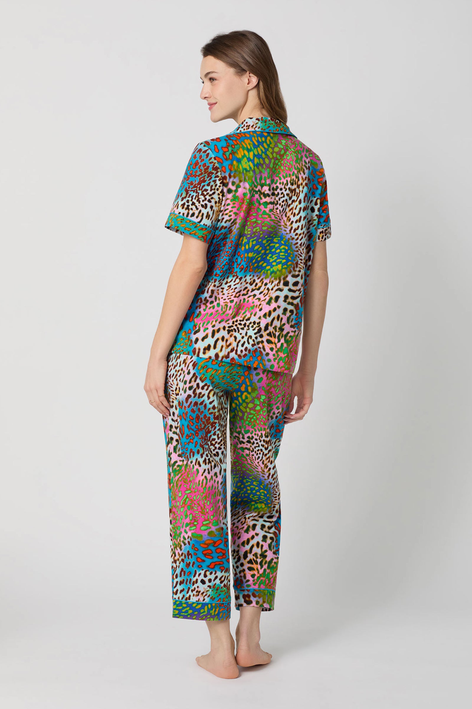A woman stands barefoot, facing away, wearing the Crop Classic PJ Set - Spray Leopard, a short-sleeve pajama set with a vibrant leopard print. The background is plain light gray.