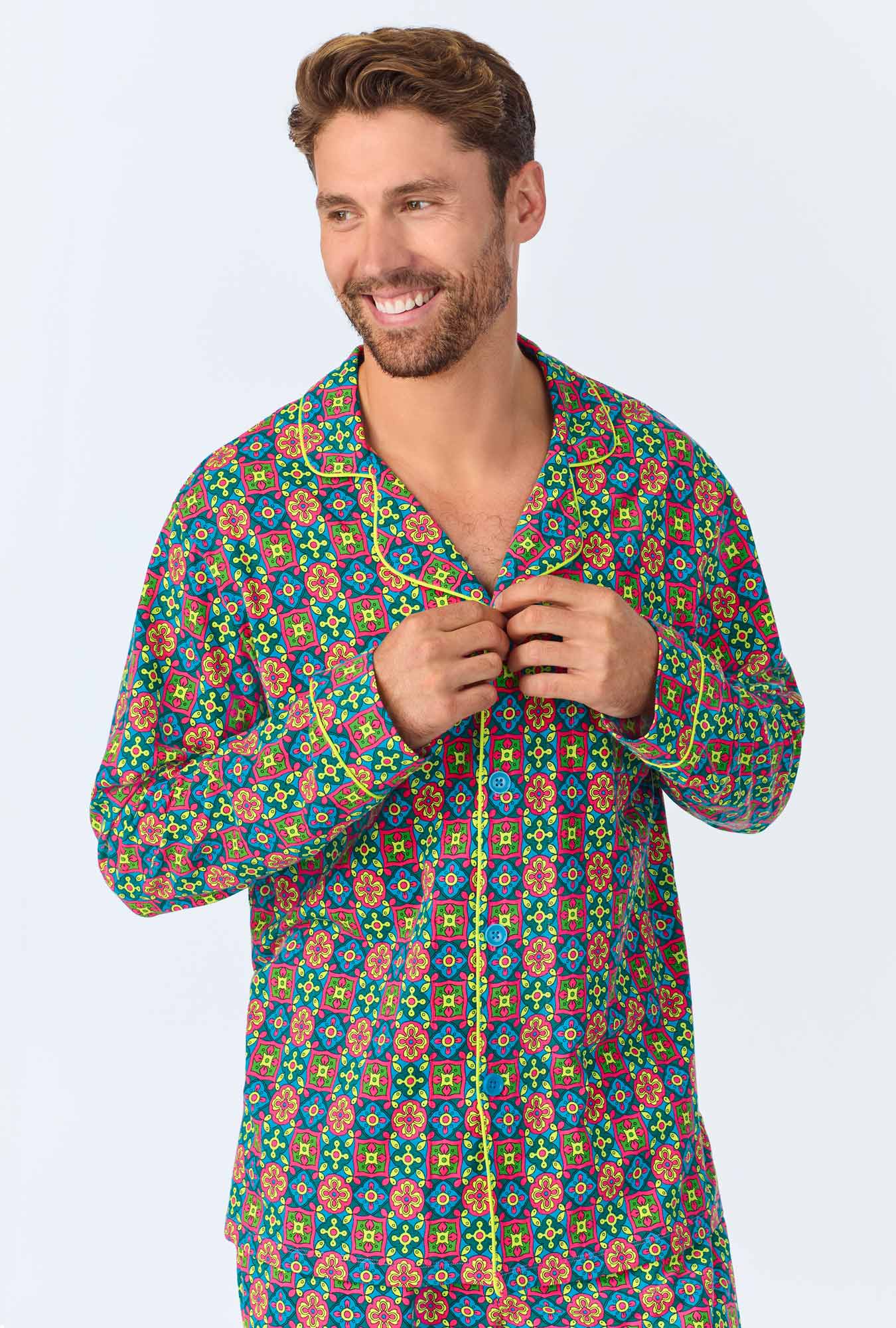 A smiling man with short brown hair buttons his shirt while wearing the Pretty Tiles Mens Long Sleeve Long Pant Jersey PJ Set, which features colorful geometric and floral designs in red, blue, yellow, and green.