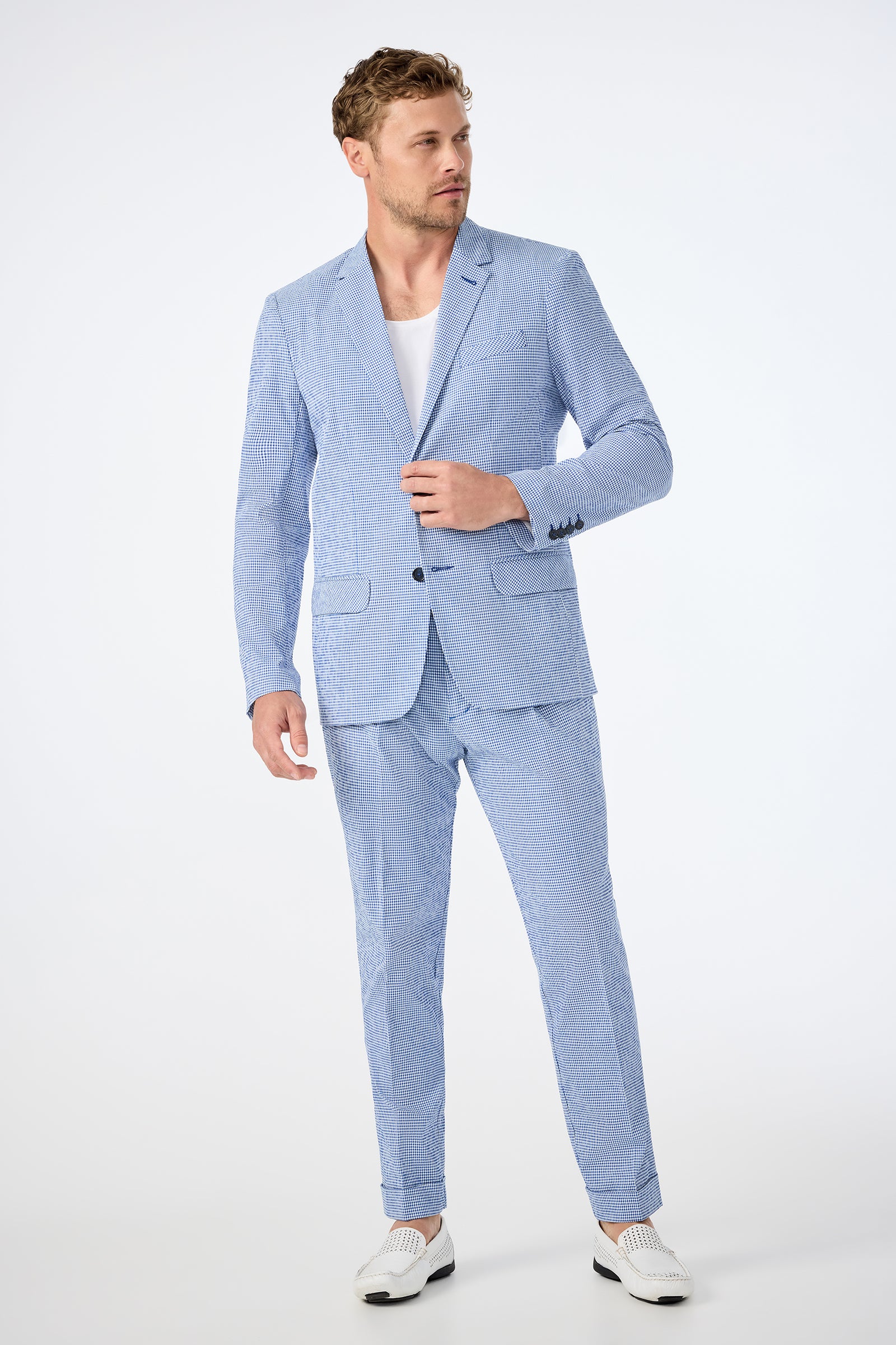 A man wears the Louie Blazer - Ocean Gingham over a white t-shirt and loafers, standing against a plain white background, looking right with one hand touching his blazer.