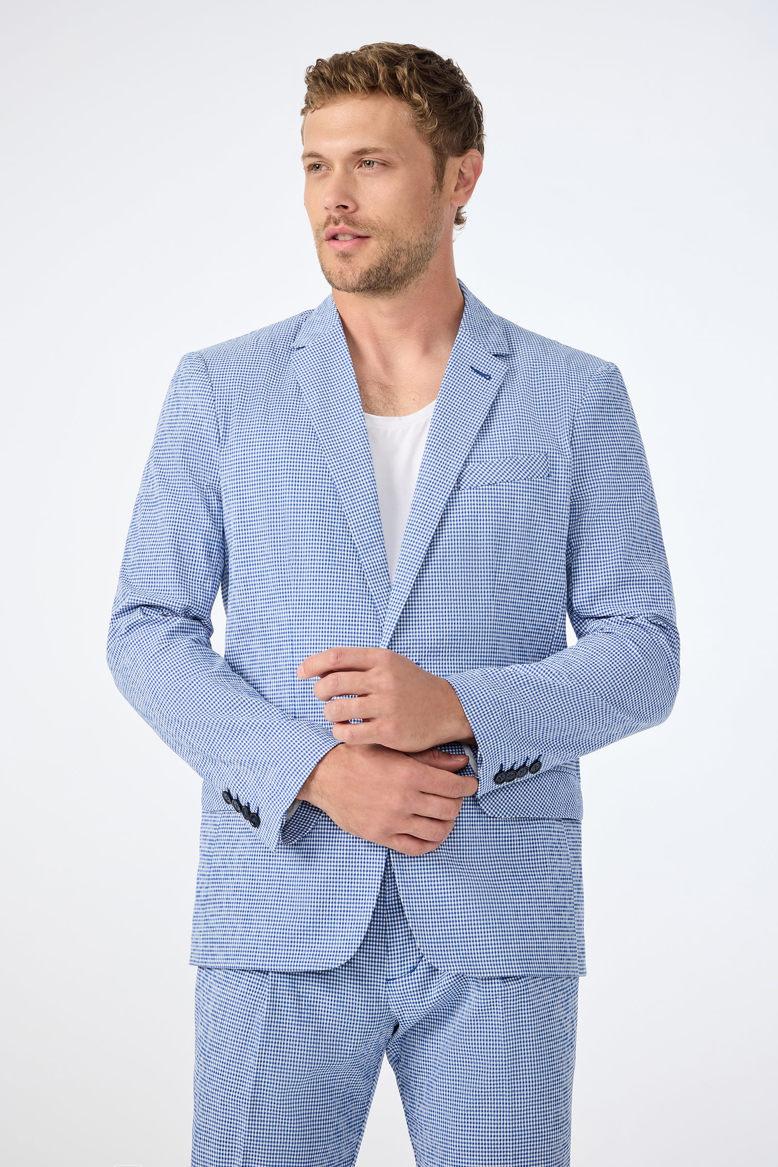 A man wears the Louie Blazer - Ocean Gingham over a white t-shirt, standing against a plain white background and adjusting his sleeve while looking slightly to the side.