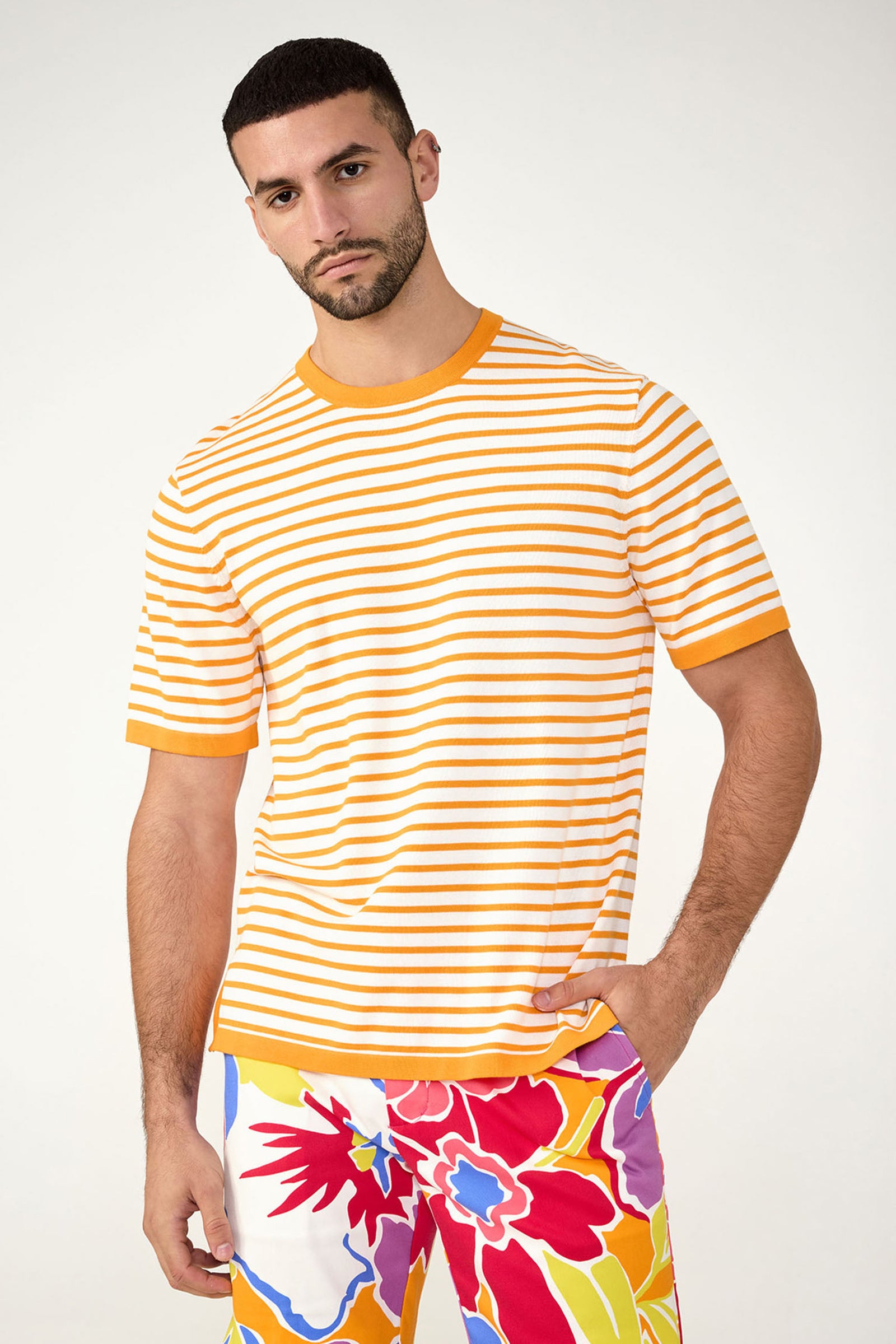 A man stands facing forward, wearing the Giovanni Stripe Short-Sleeve Pullover in Cap Clementine paired with colorful floral-patterned shorts in red, yellow, blue, and purple tones.