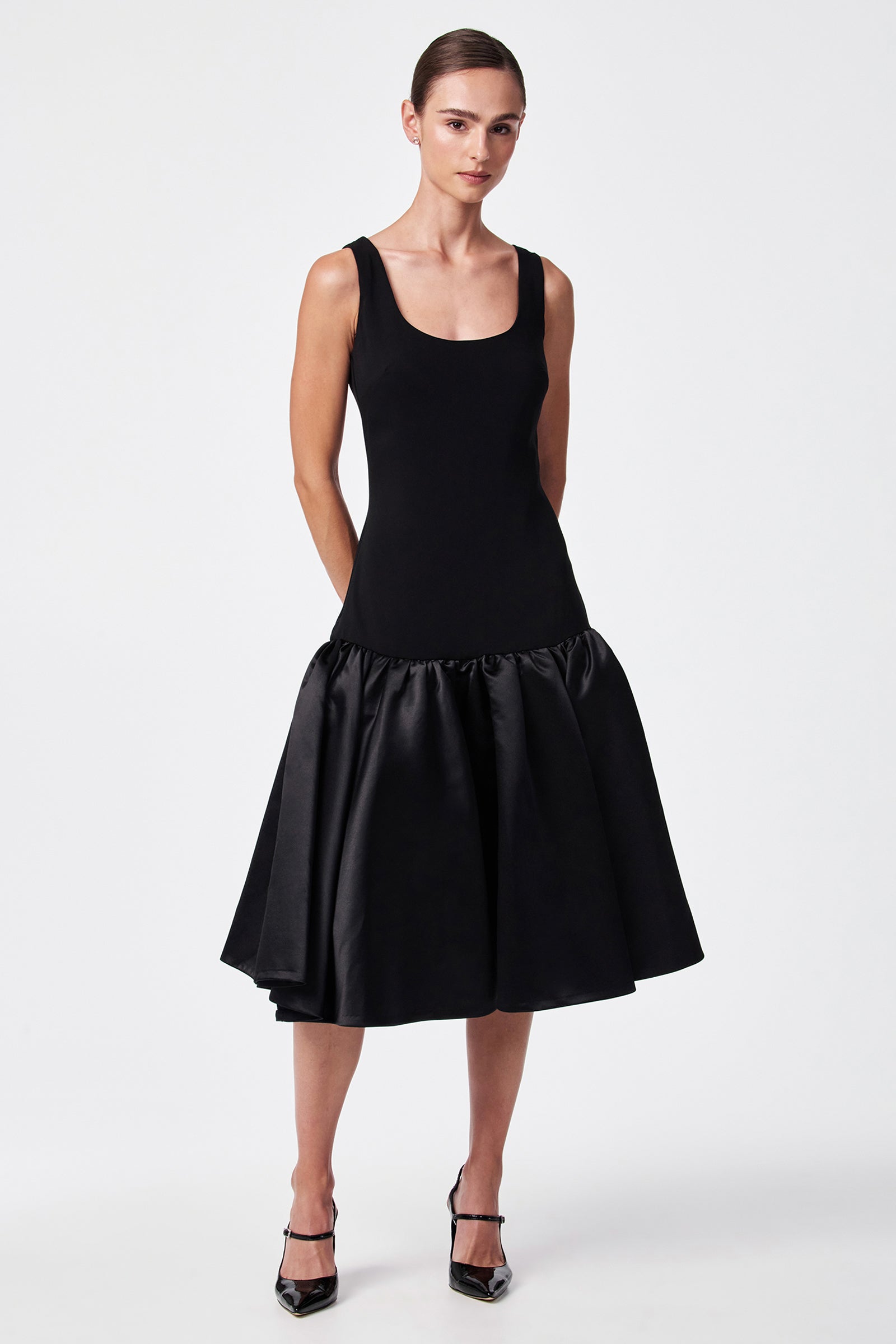 A woman stands against a white background wearing the SINA DRESS - BLACK, which features a sleeveless fitted bodice and voluminous drop-waist satin skirt, paired with black pointed-toe heels.