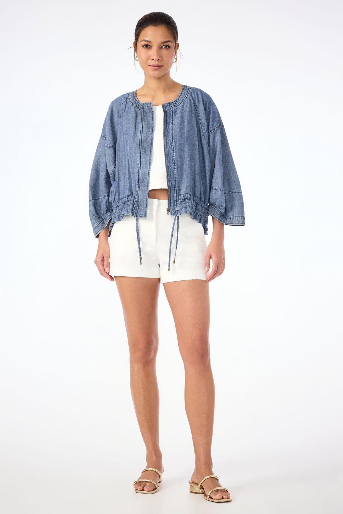 A woman stands facing forward, wearing the Prita Chambray Jacket over a white top and white shorts with sandals. Her hair is tied back, and the background is plain white.