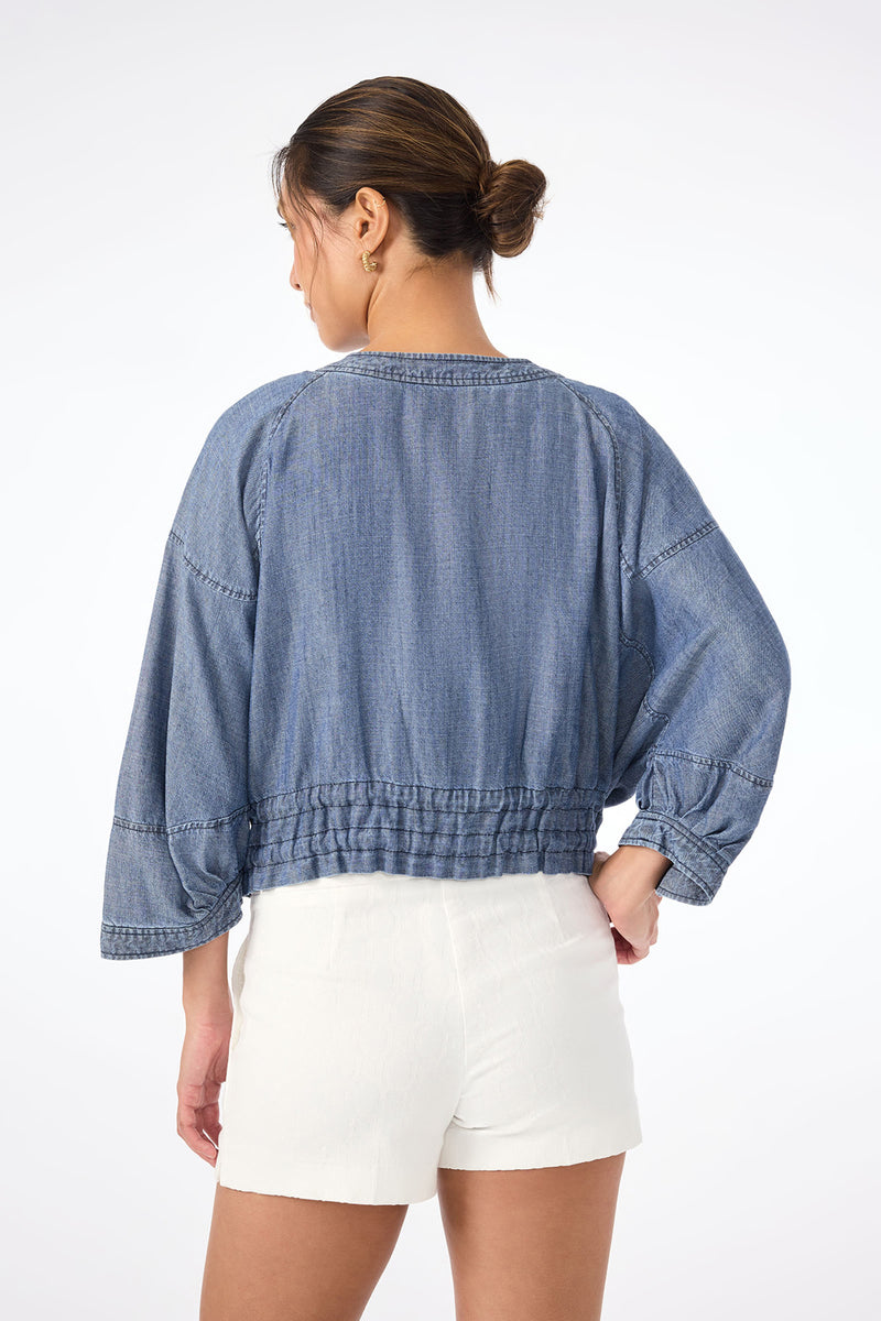 A woman with her hair in a neat bun faces away, wearing the loose-fitting Prita Chambray Jacket with gathered hem and white high-waisted shorts against a plain white background.