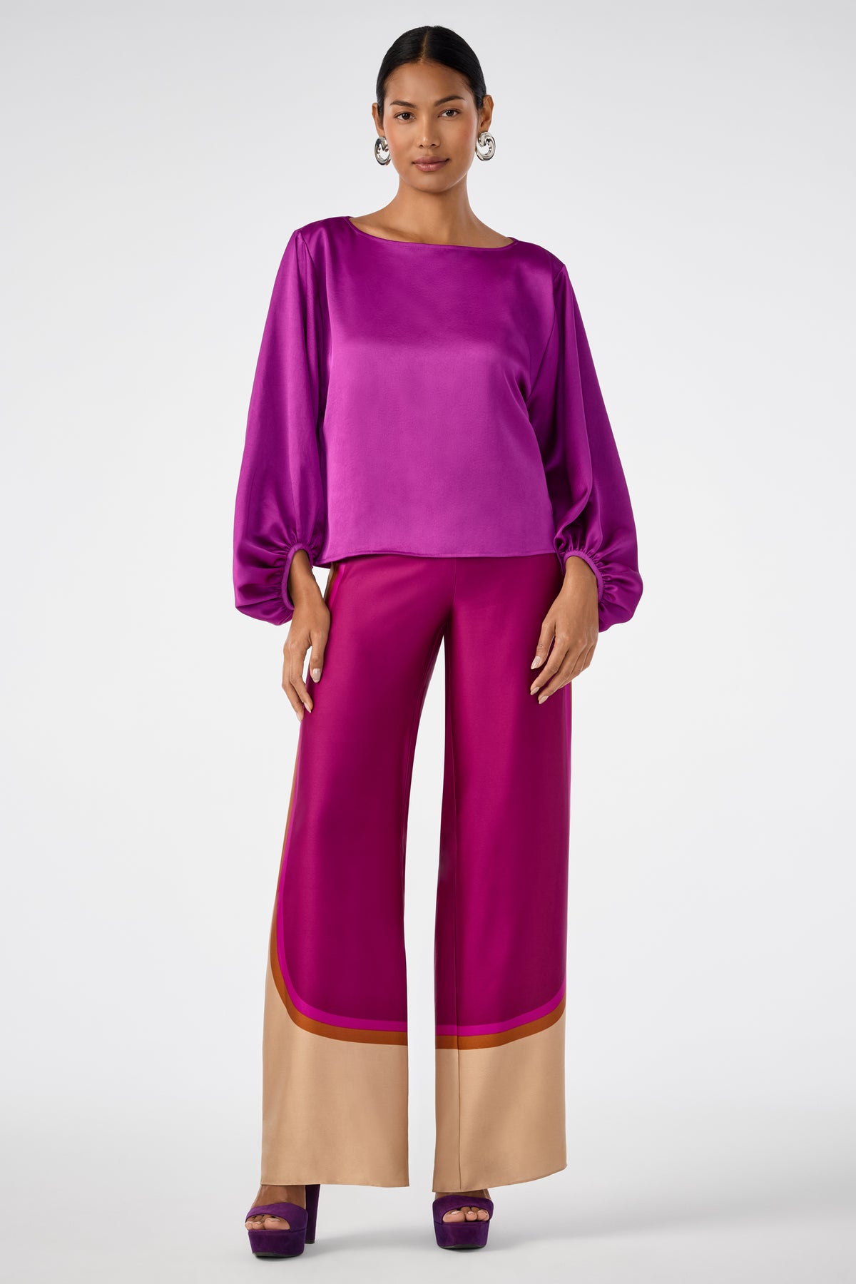 A woman stands wearing the shiny magenta Ursa 2 Top with matching wide-leg magenta pants featuring tan and red trim at the hem and purple heels, set against a plain light background.