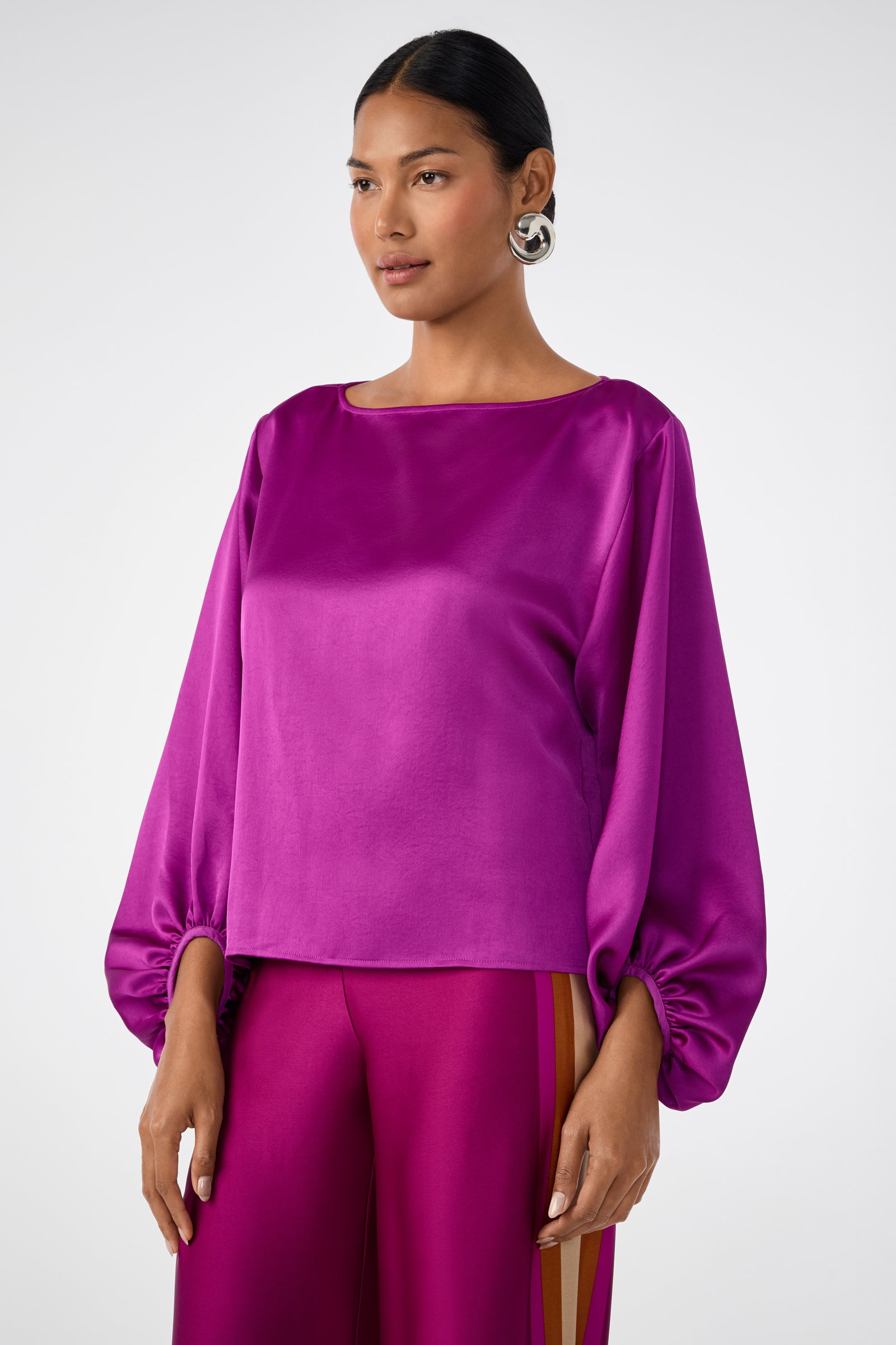 A woman wears the Ursa 2 Top, a shiny long-sleeved magenta blouse with ruched cuffs, paired with matching magenta pants featuring yellow side stripes, standing against a plain light background.