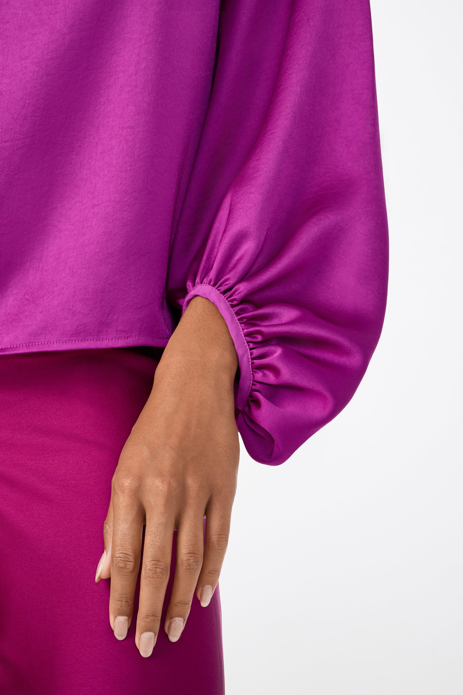 A person wears the Ursa 2 Top, a bright magenta long-sleeve with gathered cuffs, paired with matching pants. Only their waist down and one hand are shown.