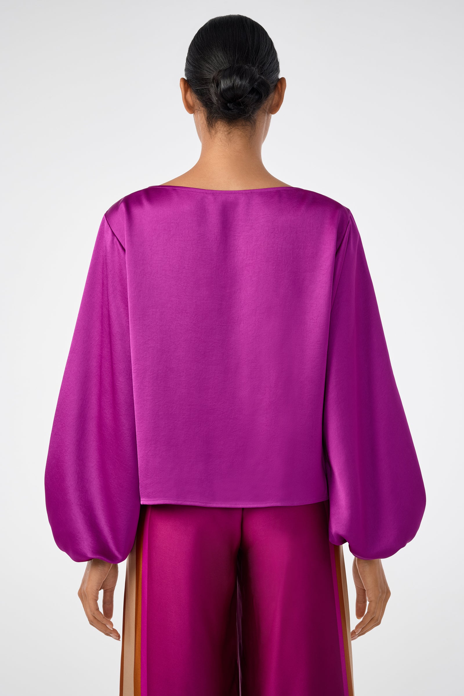 A person with dark hair in a low bun is shown from the back, wearing the shiny magenta Ursa 2 Top with long, loose sleeves and matching magenta pants with beige side stripes against a plain, light background.
