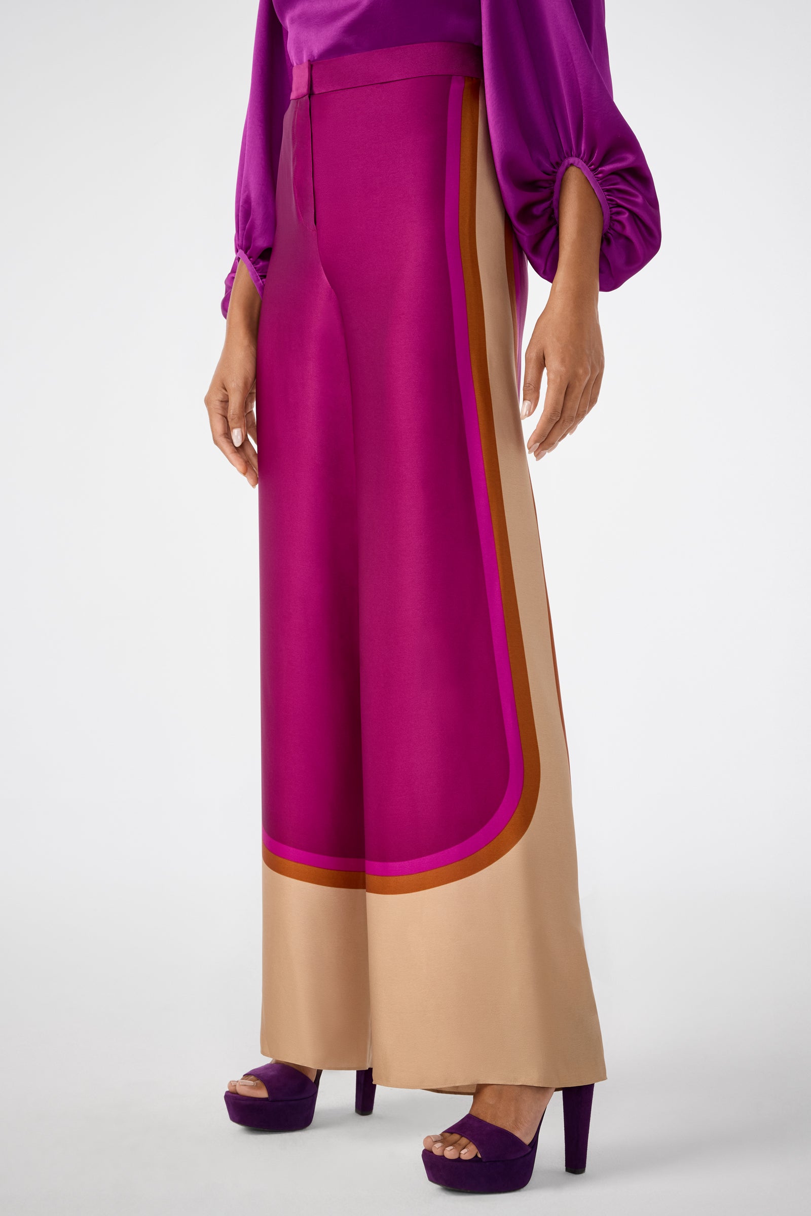 A person wears the Verity 2 Pant—wide-leg, high-waisted with bold magenta, tan, and orange color blocking—paired with a purple top and matching platform heels.