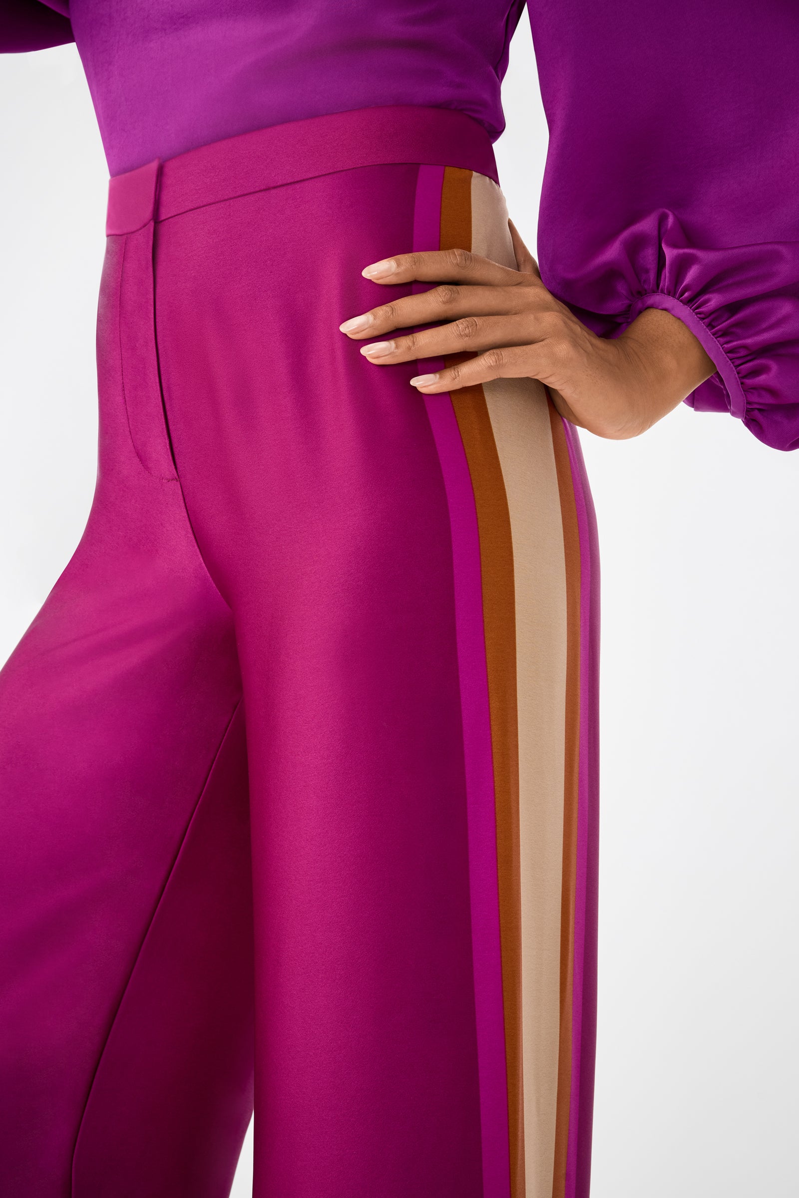 A person wearing the Verity 2 Pant in magenta satin with mustard, beige, and pink side stripes, paired with a matching puff-sleeve magenta top, stands with one hand on their hip.