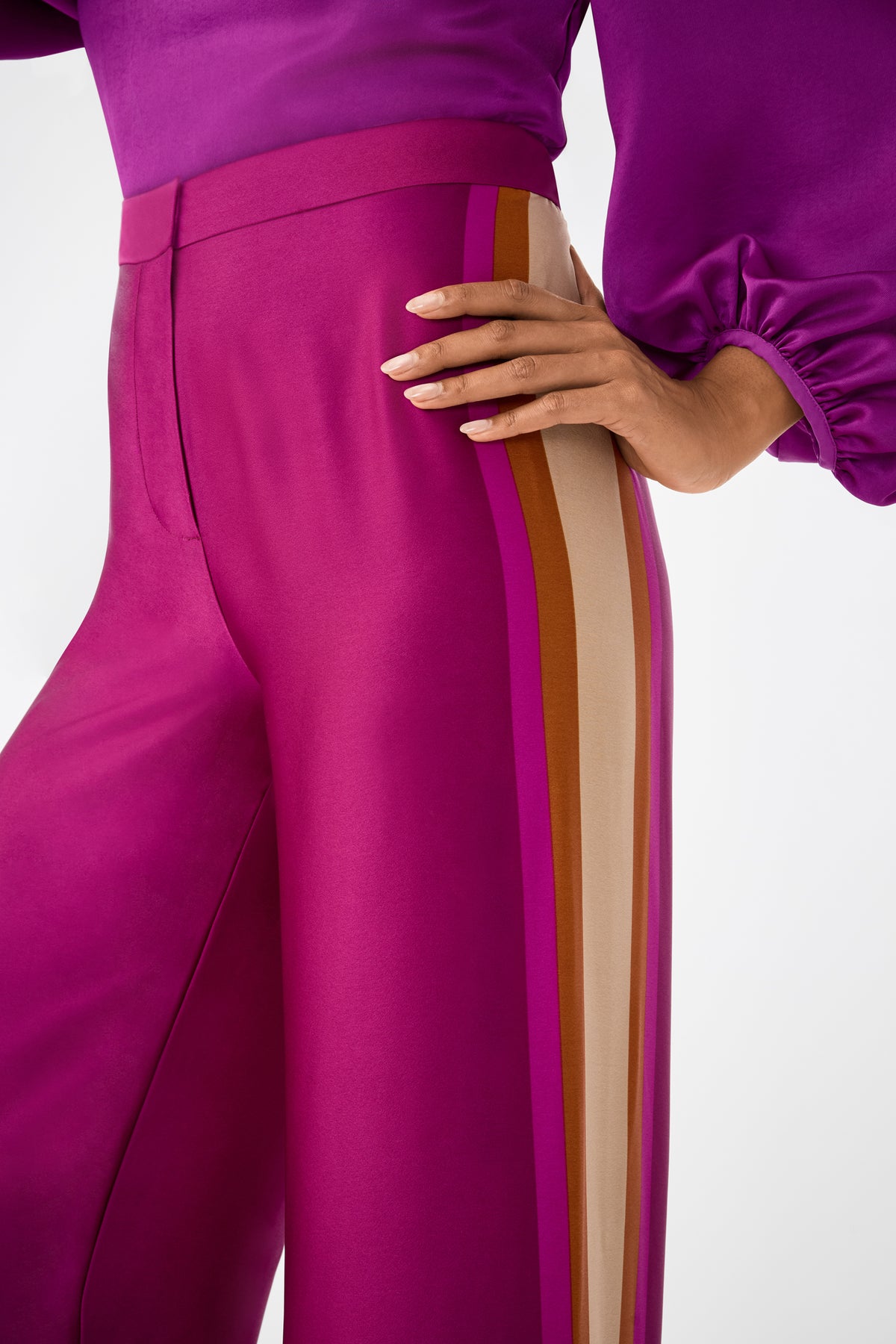 A person wearing the Verity 2 Pant in magenta satin with mustard, beige, and pink side stripes, paired with a matching puff-sleeve magenta top, stands with one hand on their hip.