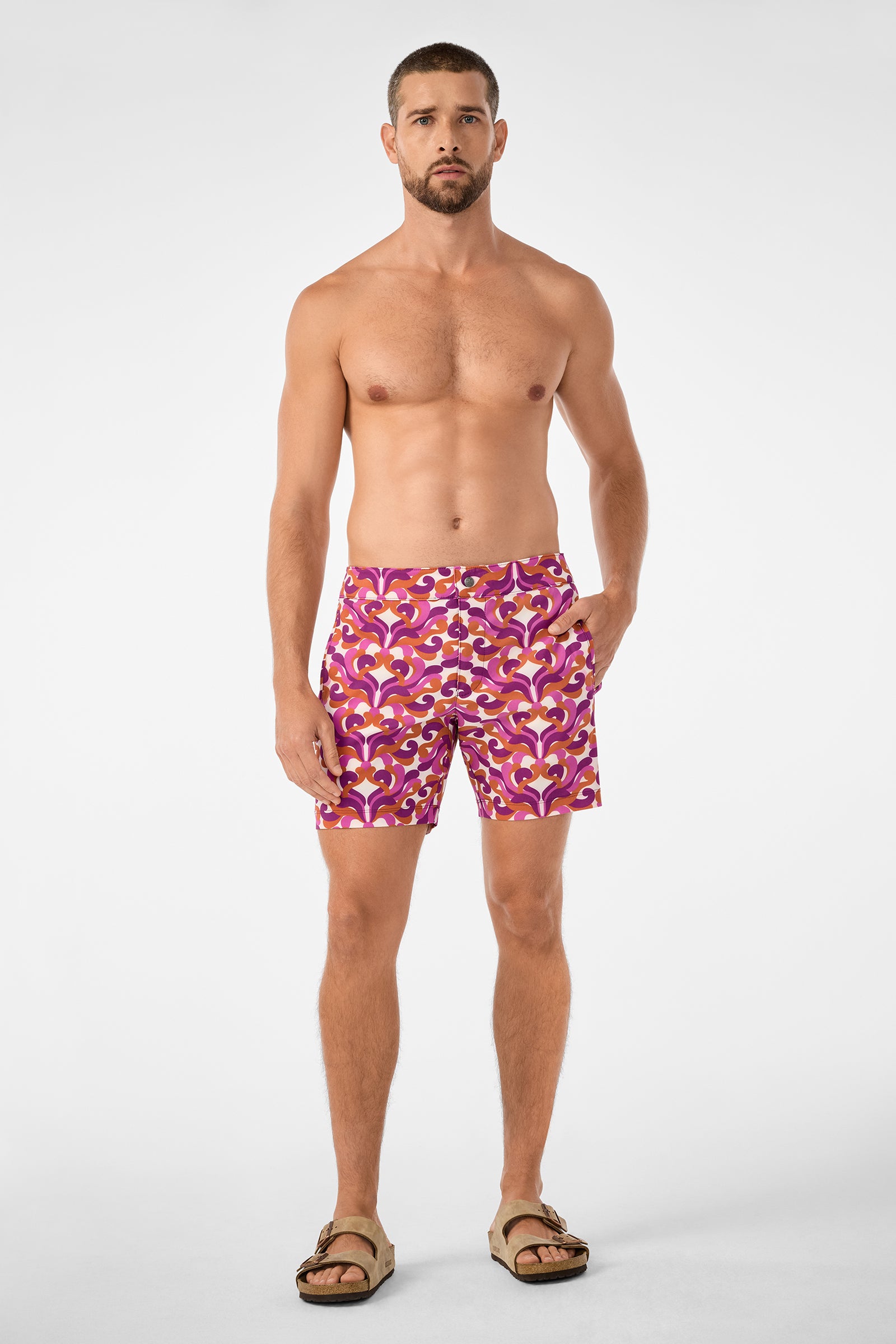 A man with a short beard stands barefoot against a white background, wearing the Honolulu Board Short in a pink, red, and purple pattern with tan sandals. He is shirtless and looks straight ahead with a neutral expression.