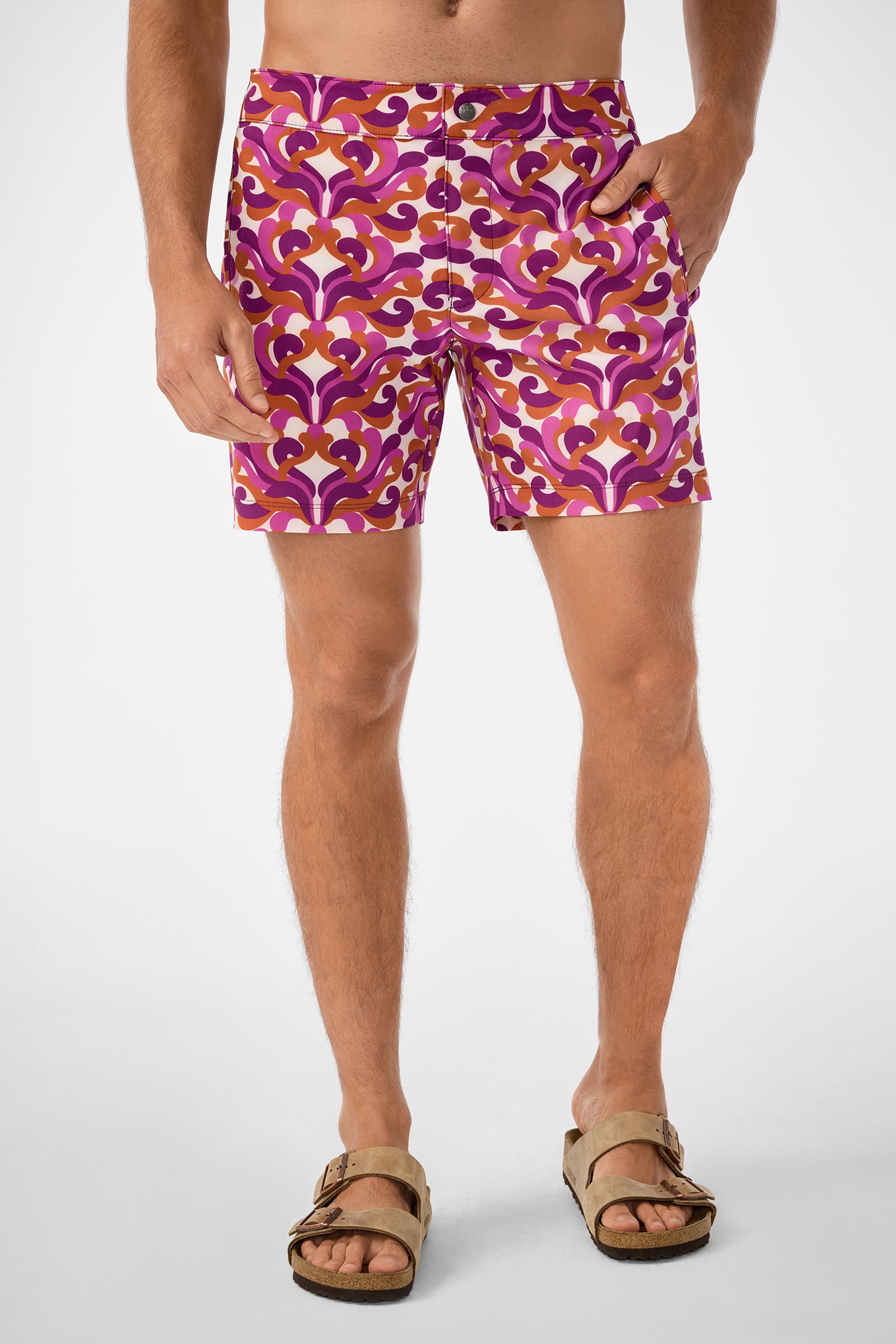 A person models the Honolulu Board Short—colorful retro-patterned swim shorts in purple, pink, and orange—with tan sandals. The cropped image (chest to feet) features a plain white background.
