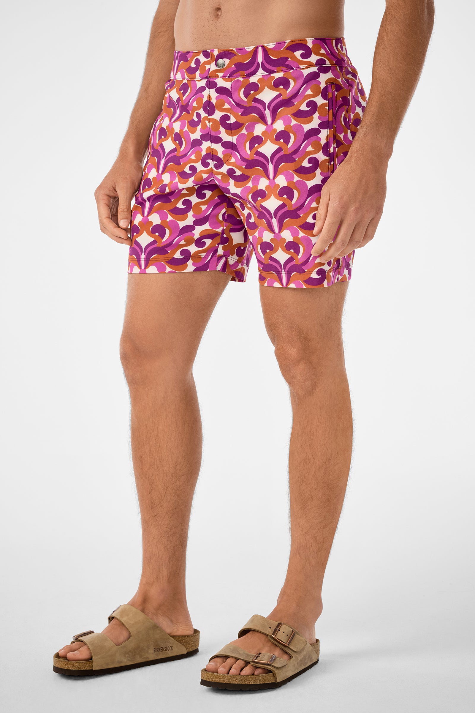 A person models the Honolulu Board Short featuring vibrant purple, pink, and orange retro patterns, paired with tan sandals. The image is cropped from shoulders down against a plain light background.