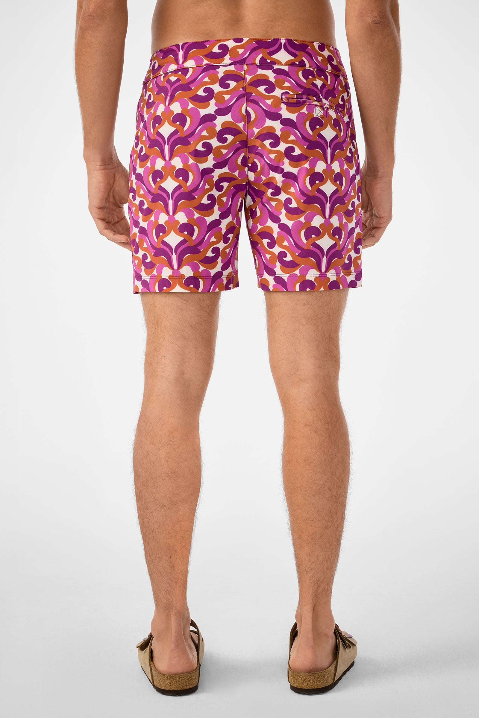 A man faces away in Honolulu Board Short swim trunks featuring a retro pink, orange, and purple pattern. He wears brown sandals, set against a plain white background.
