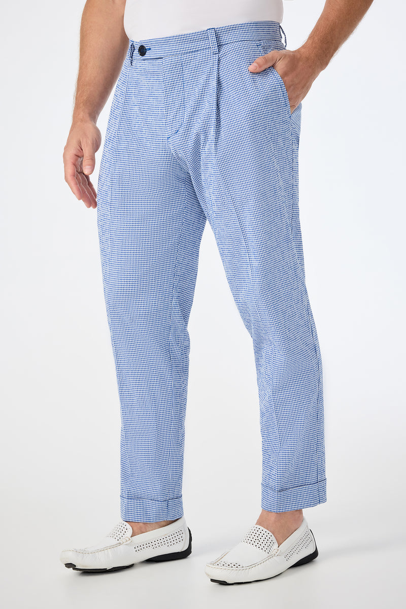 Wearing the Beale Pleated Trouser in Ocean Gingham, a man pairs these light blue and white checkered pants with a white shirt and woven loafers. With his left hand in his pocket, only his body from the shoulders down is shown on a white background.