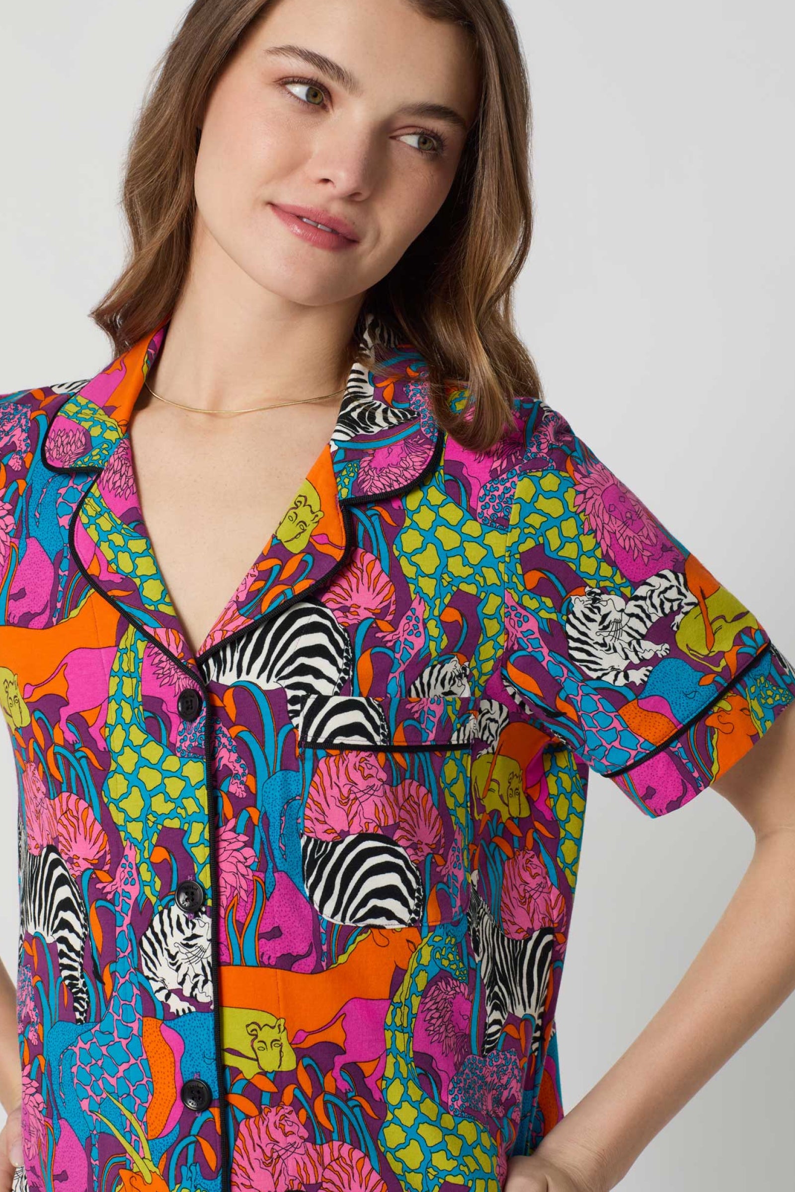 A woman wears the Shorty PJ Set - Jungle, a vibrant short-sleeve button-up with a bold multicolored pattern of zebras, leaves, and abstract shapes. She gazes to the side with a soft smile.