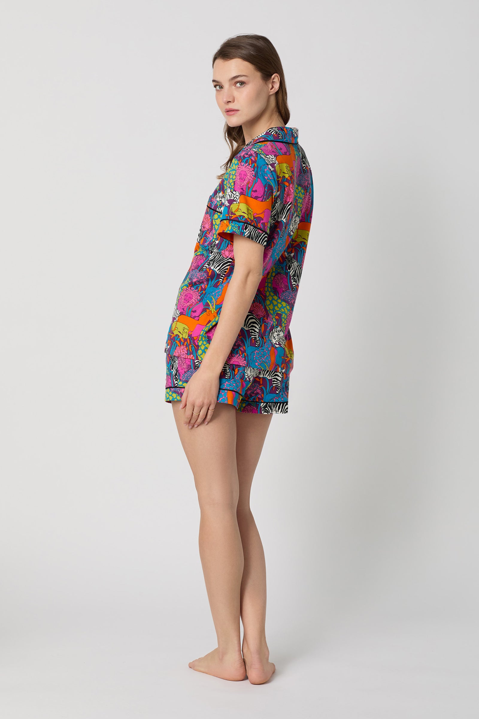 A woman stands barefoot in profile, wearing the Shorty PJ Set - Jungle, featuring a vibrant, colorful animal print with a matching short-sleeve shirt and shorts. She faces left against a plain white background.