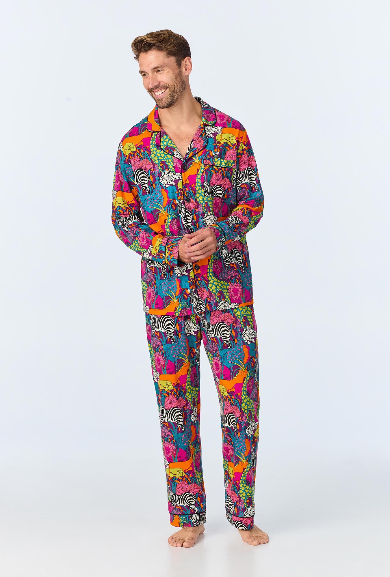 A man stands barefoot, smiling and wearing the Jungle Mens Long Sleeve Long Pant Jersey PJ Set in multi, featuring a bold animal print with zebras and lions on a white background.