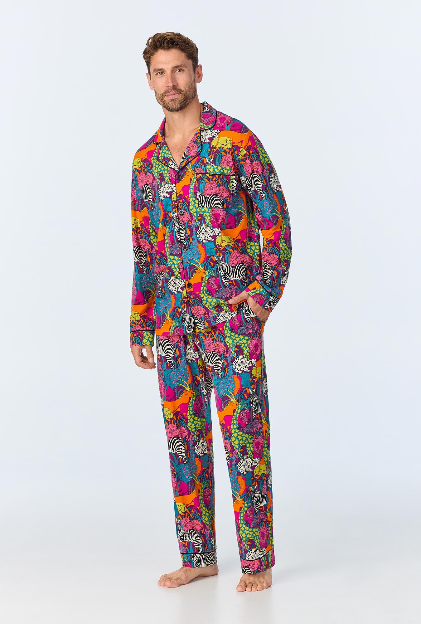 A man with short hair and a beard stands barefoot in the Jungle Mens Long Sleeve Long Pant Jersey PJ Set, featuring a vibrant animal print with zebras and other creatures on a white stretch jersey background. He looks relaxed.