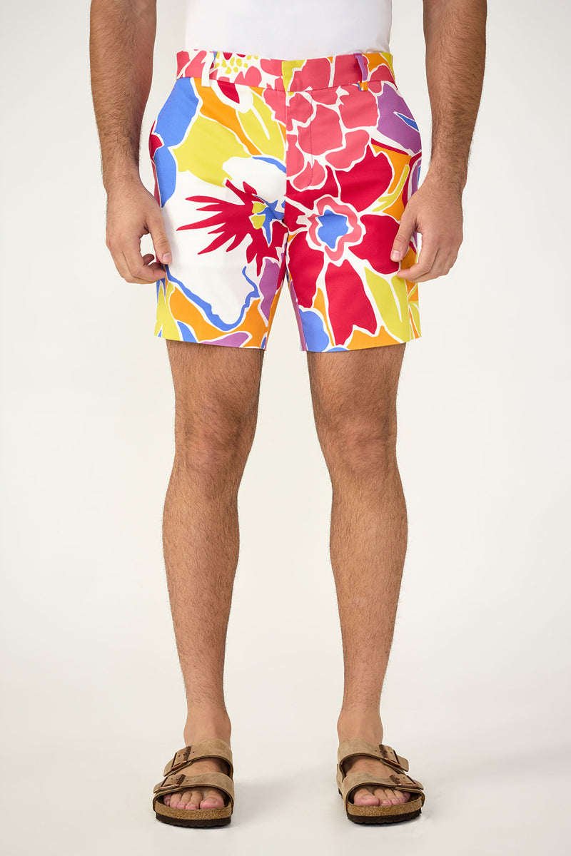 Model wears the Lawrence Short - Soi Paradis in MULTI, featuring red, yellow, blue, and white abstract floral patterns, paired with brown sandals. Shown from waist to feet against a plain light background.