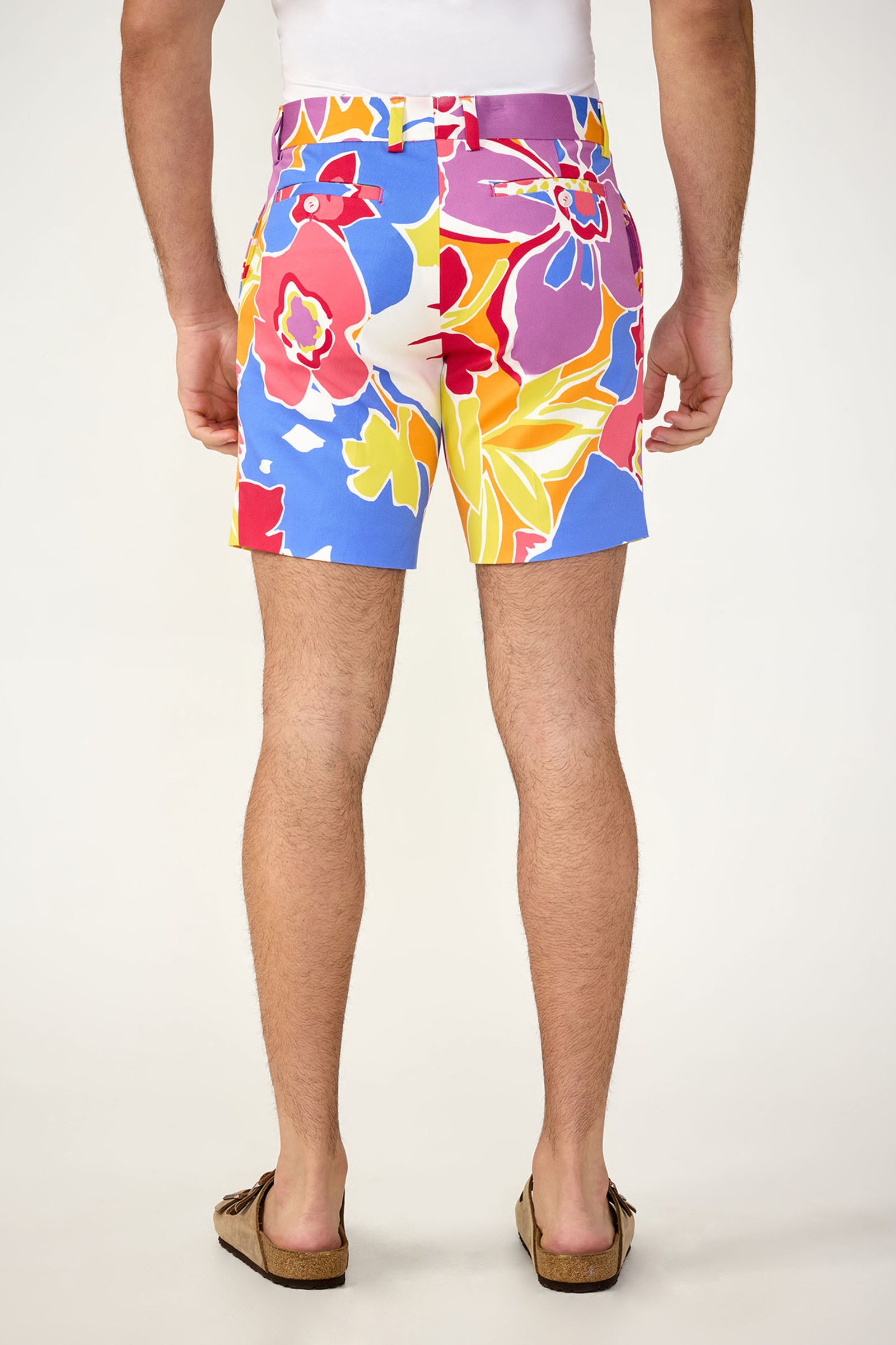 A person seen from behind wearing the Lawrence Short - Soi Paradis, featuring bright multicolored florals in red, yellow, pink, and blue, paired with brown sandals and a white top.