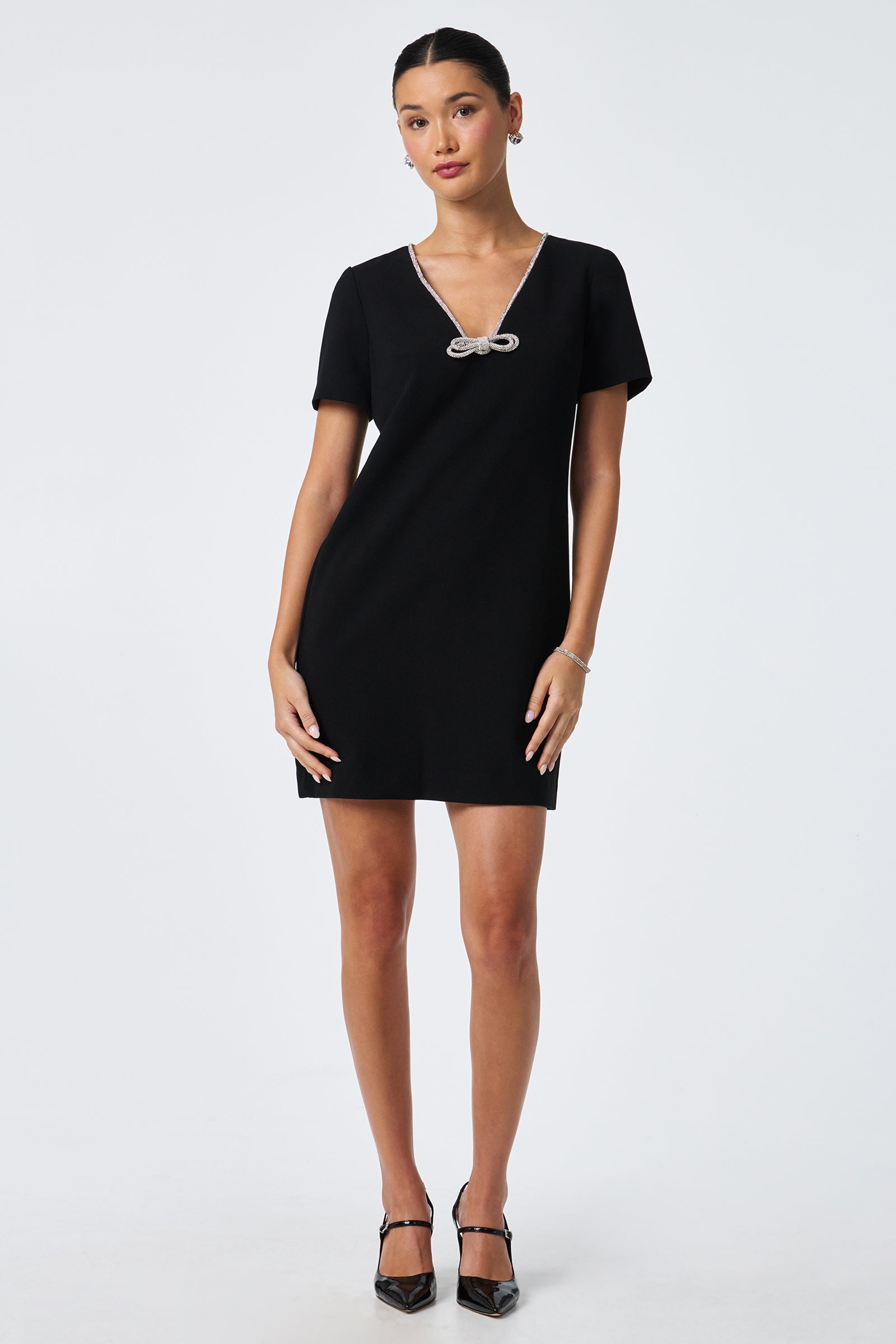 A woman stands against a white background wearing the Obelle Crystal Trim Mini Dress, featuring short sleeves, a deep V-neckline with a decorative bow, and black heels. Her hair is tied back and her arms are relaxed at her sides.