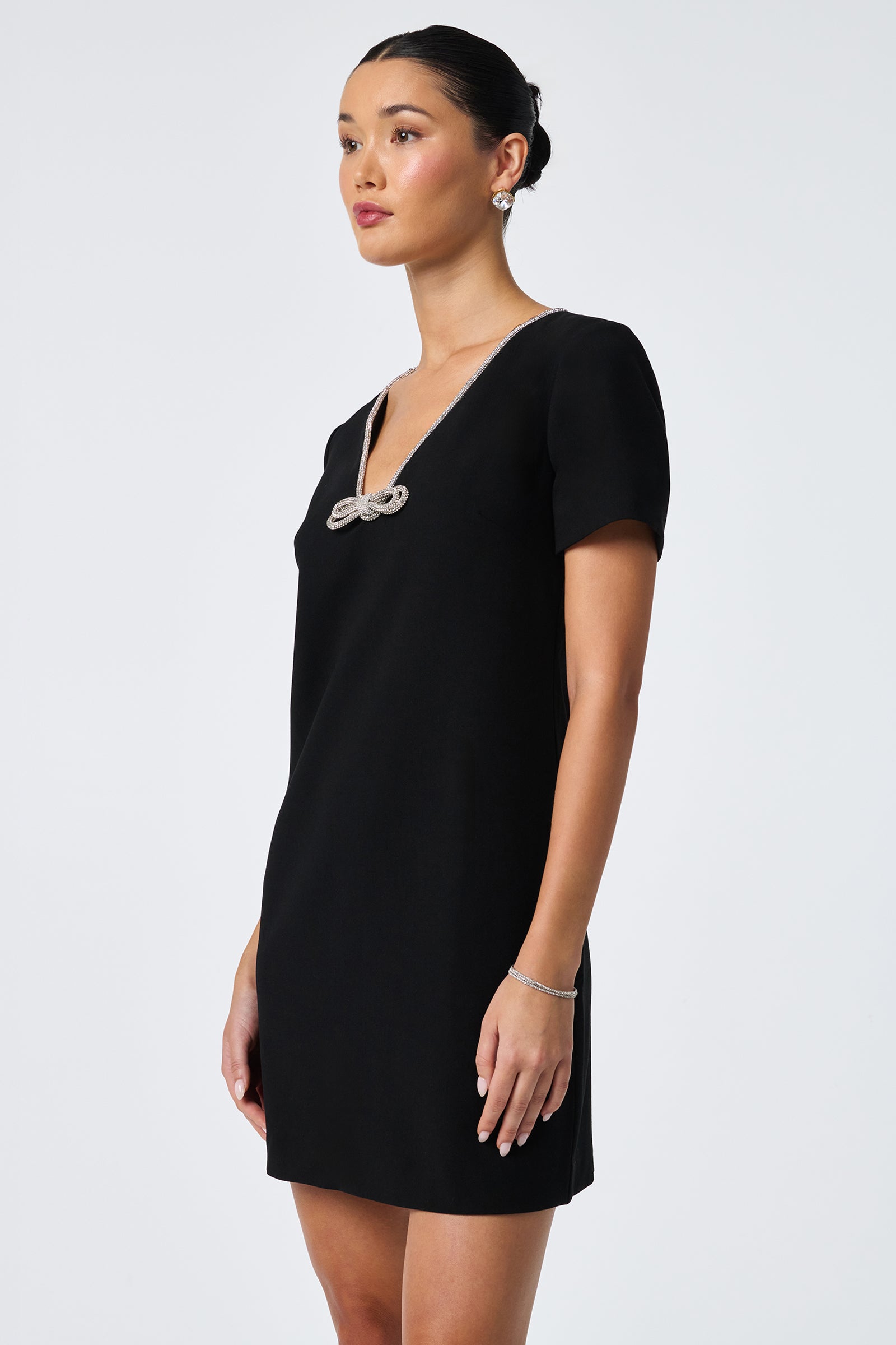 A woman models the Obelle Crystal Trim Mini Dress, featuring short sleeves, a V-neckline with sparkling crystal trim, and a chic black mini silhouette against a plain light background.