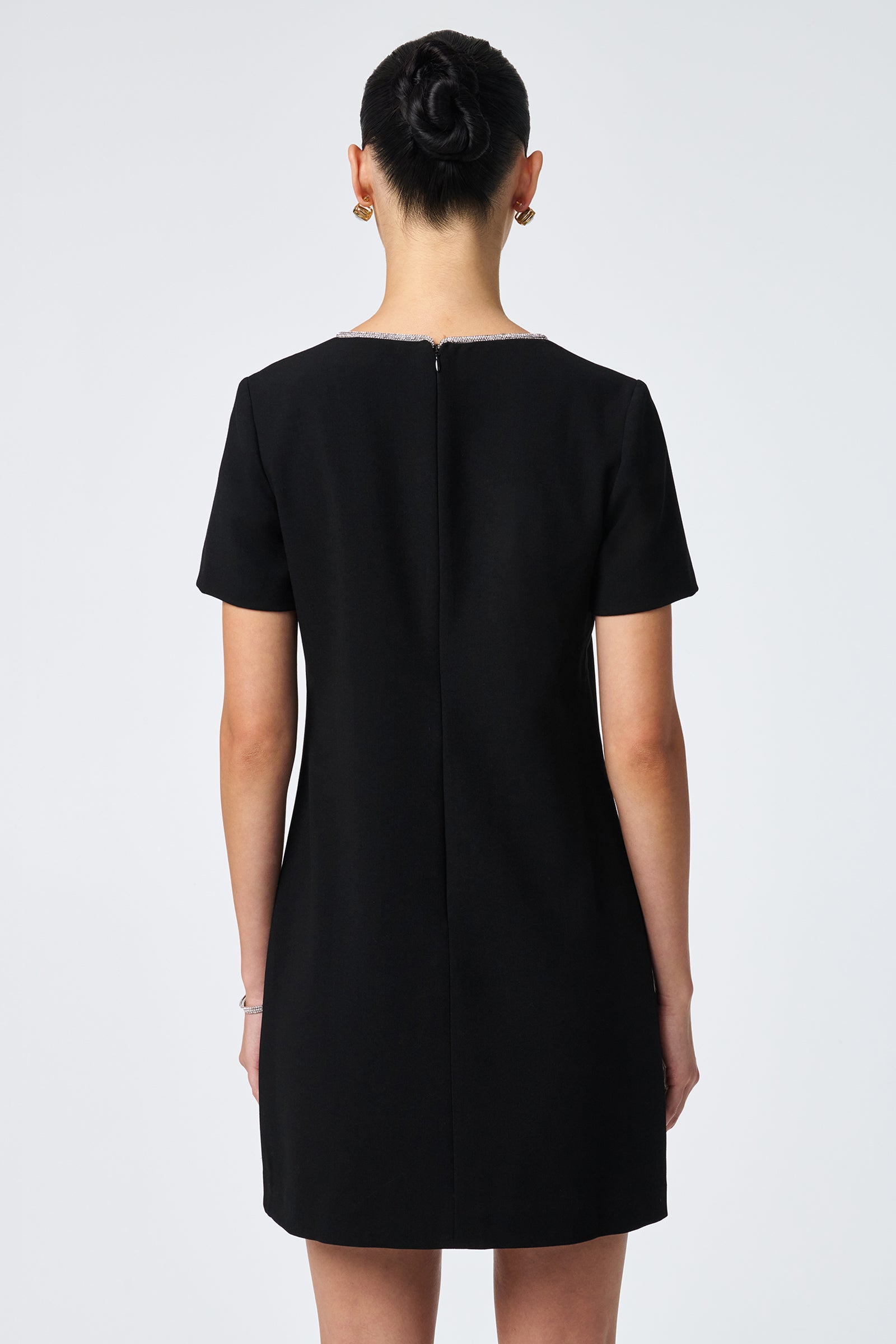 A woman with dark hair in a neat bun is shown from the back, wearing the Obelle Crystal Trim Mini Dress. She stands against a plain, light background.